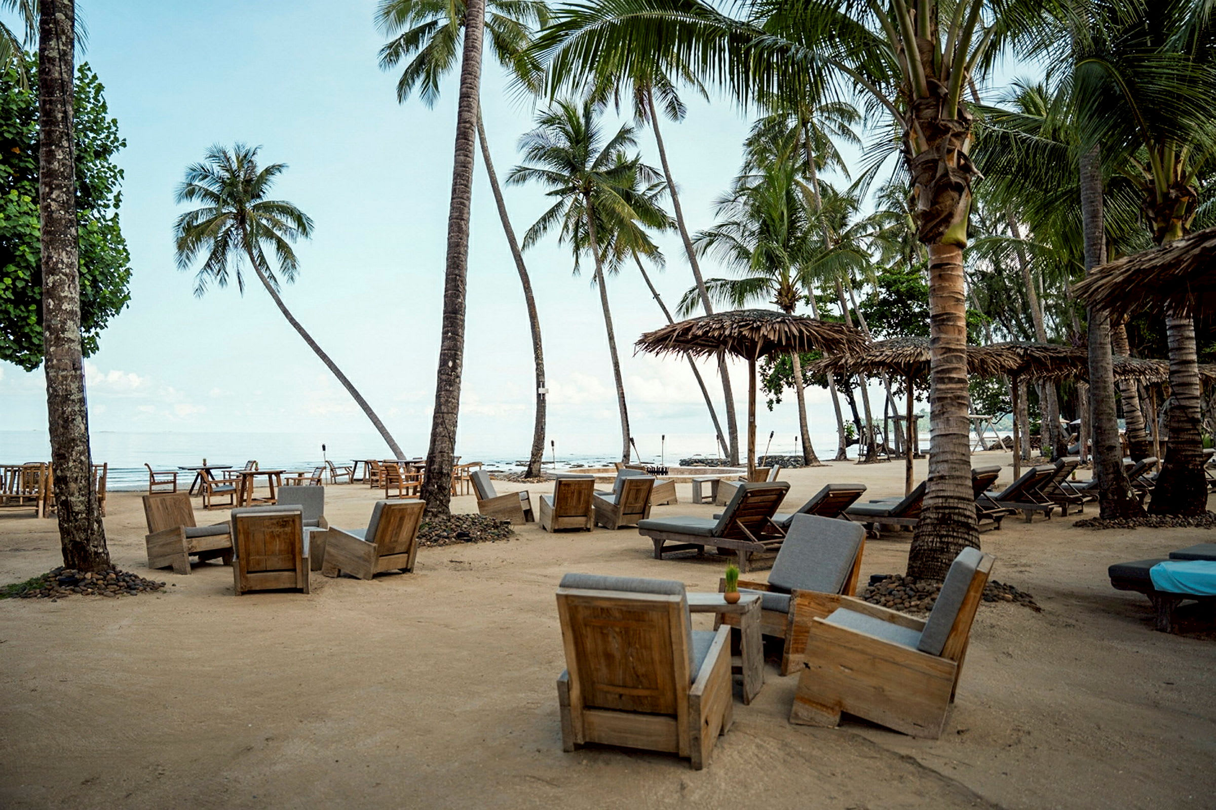 Strand med solstolar och sittgrupper i trä, omgiven av höga palmträd. (AI-genererad text)