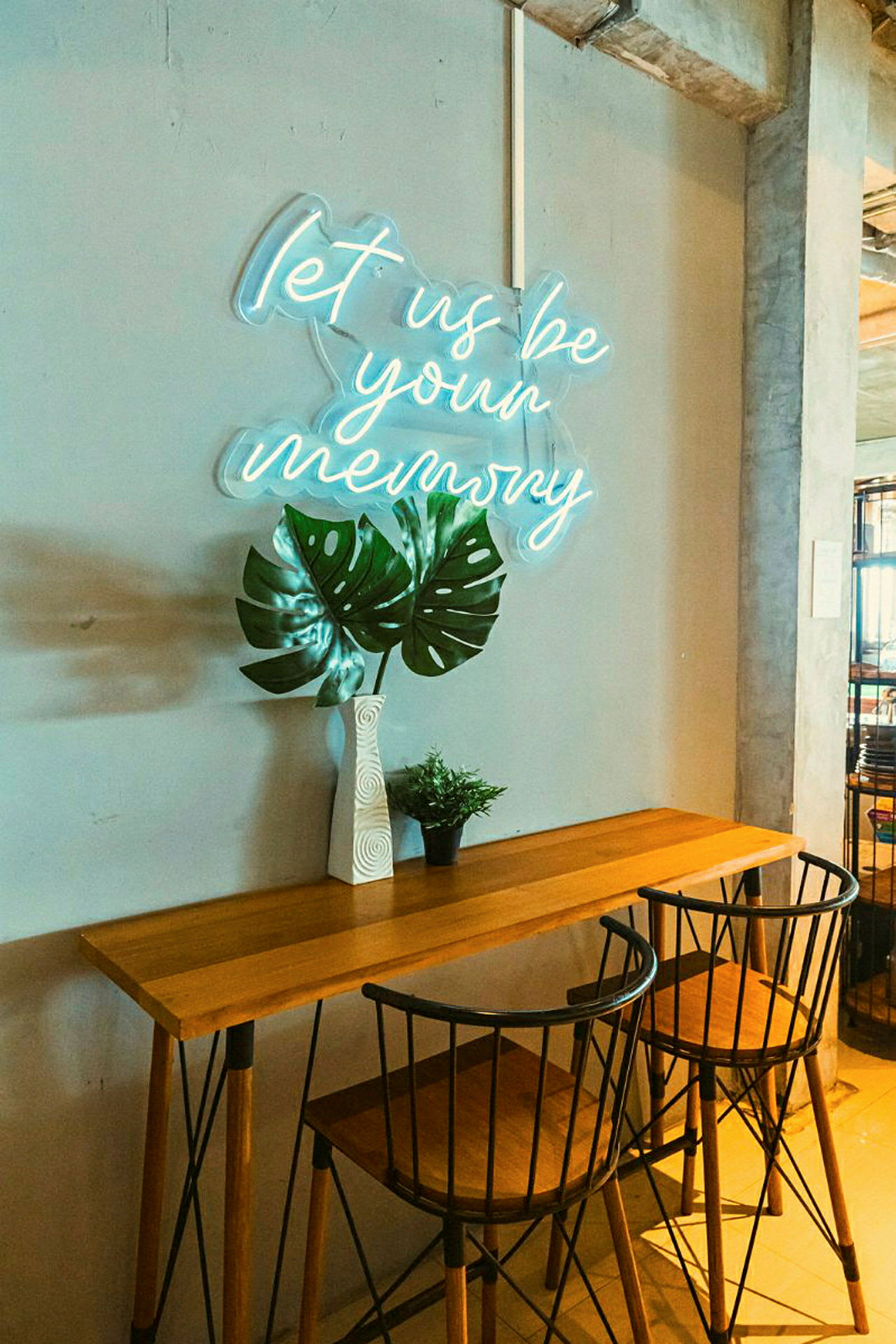 Ett bord med två stolar, en vas med stora gröna blad och en liten grön växt, framför en vägg med en neonskylt som lyser upp texten 'let us be your memory'. (AI-genererad text)