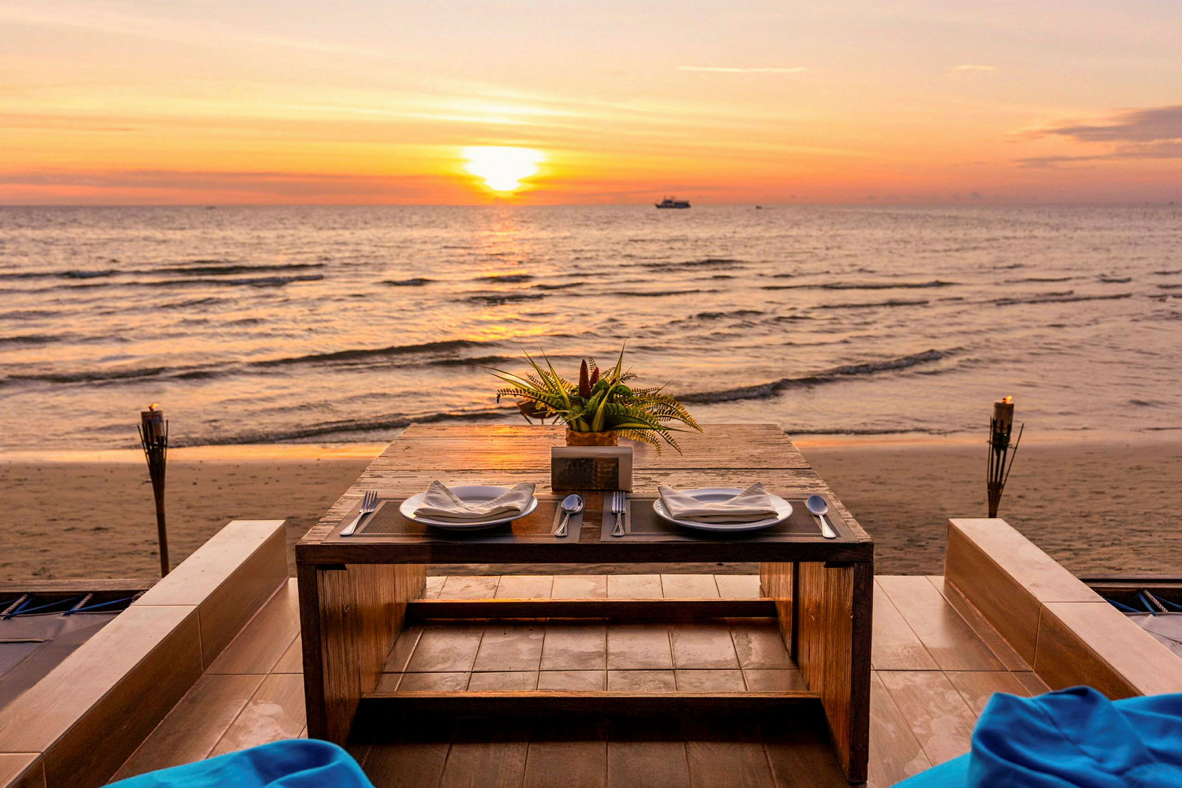 Ett dukat bord med två tallrikar och bestick står vid havet under en solnedgång. (AI-genererad text)
