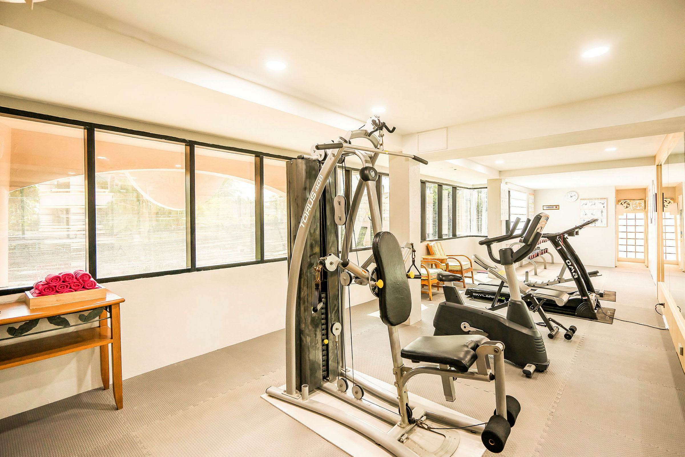 Ett välbelyst gym med träningsmaskiner, fönster och en bänk med handdukar. (AI-genererad text)