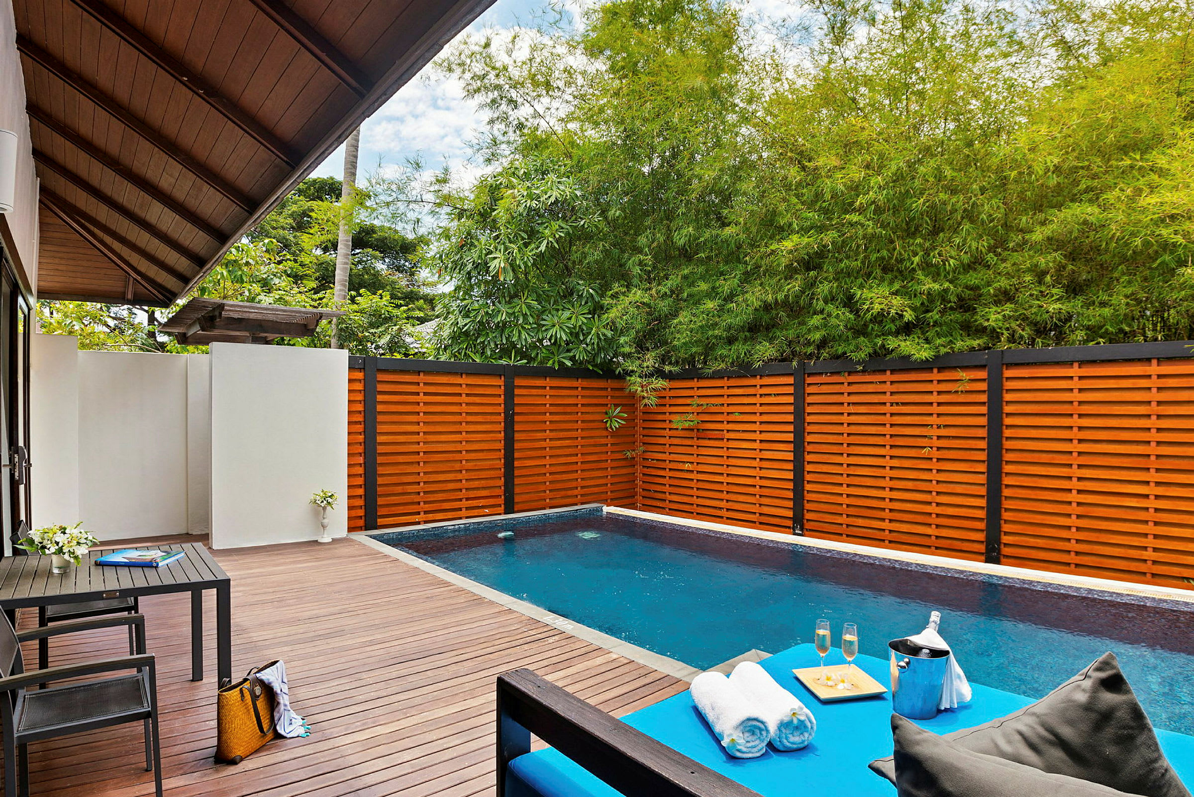 En privat pool med trädäck, omgiven av trästaket och grönska. En lounge med handdukar, drycker och en kaffebricka syns i förgrunden. (AI-genererad text)