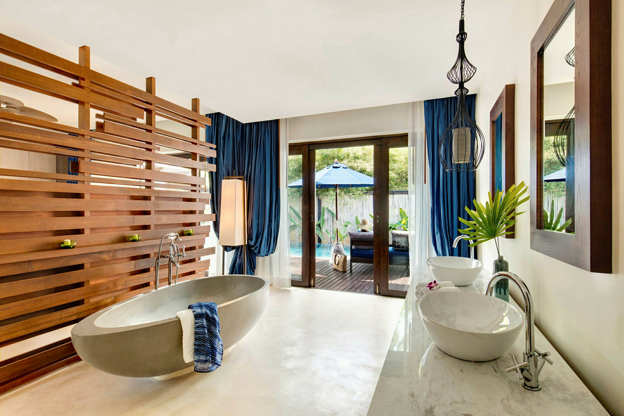 En lyxig badrum med moderna möbler, inklusive ett fristående badkar med handdukar bredvid, och en avdelare av trä som skapar en elegant känsla. Genom glasdörrar syns en privat poolområde med parasoll och solstolar. (AI-genererad text)