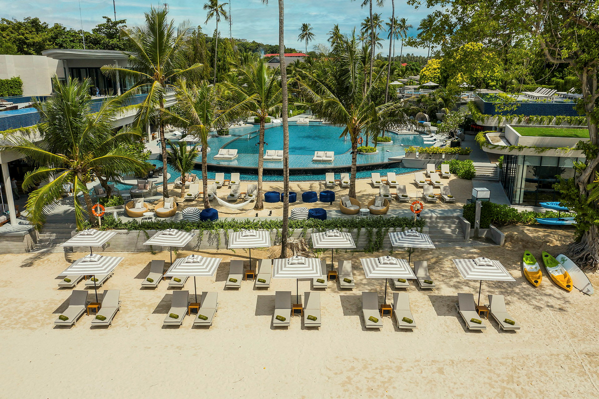 En sandstrand med solstolar och parasoller framför en lyxig pool omgiven av palmer och byggnader. (AI-genererad text)