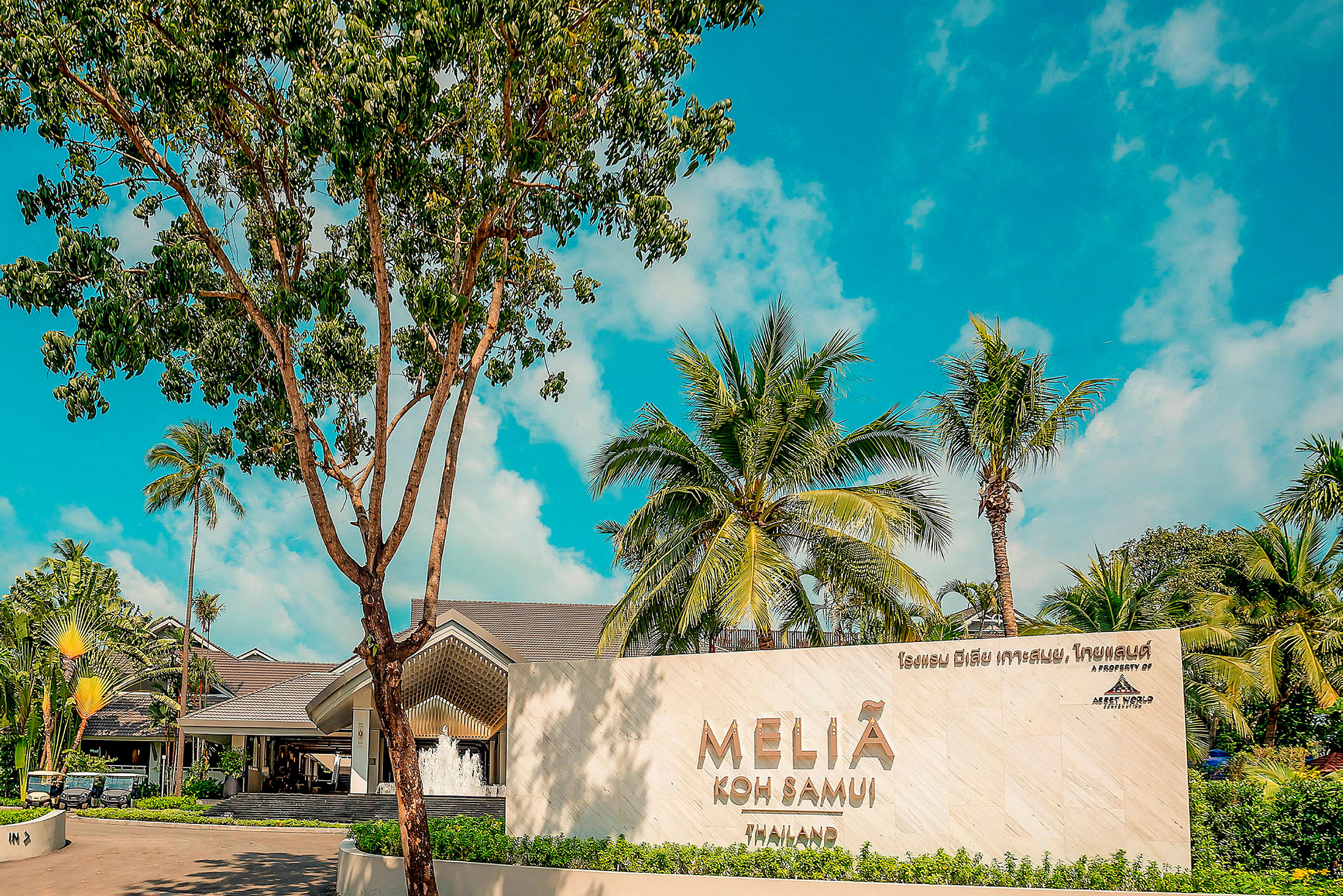 En tropisk resortinträde med en skylt som säger "Melia Koh Samui", omgiven av palmer och grönska med en blå himmel i bakgrunden. (AI-genererad text)