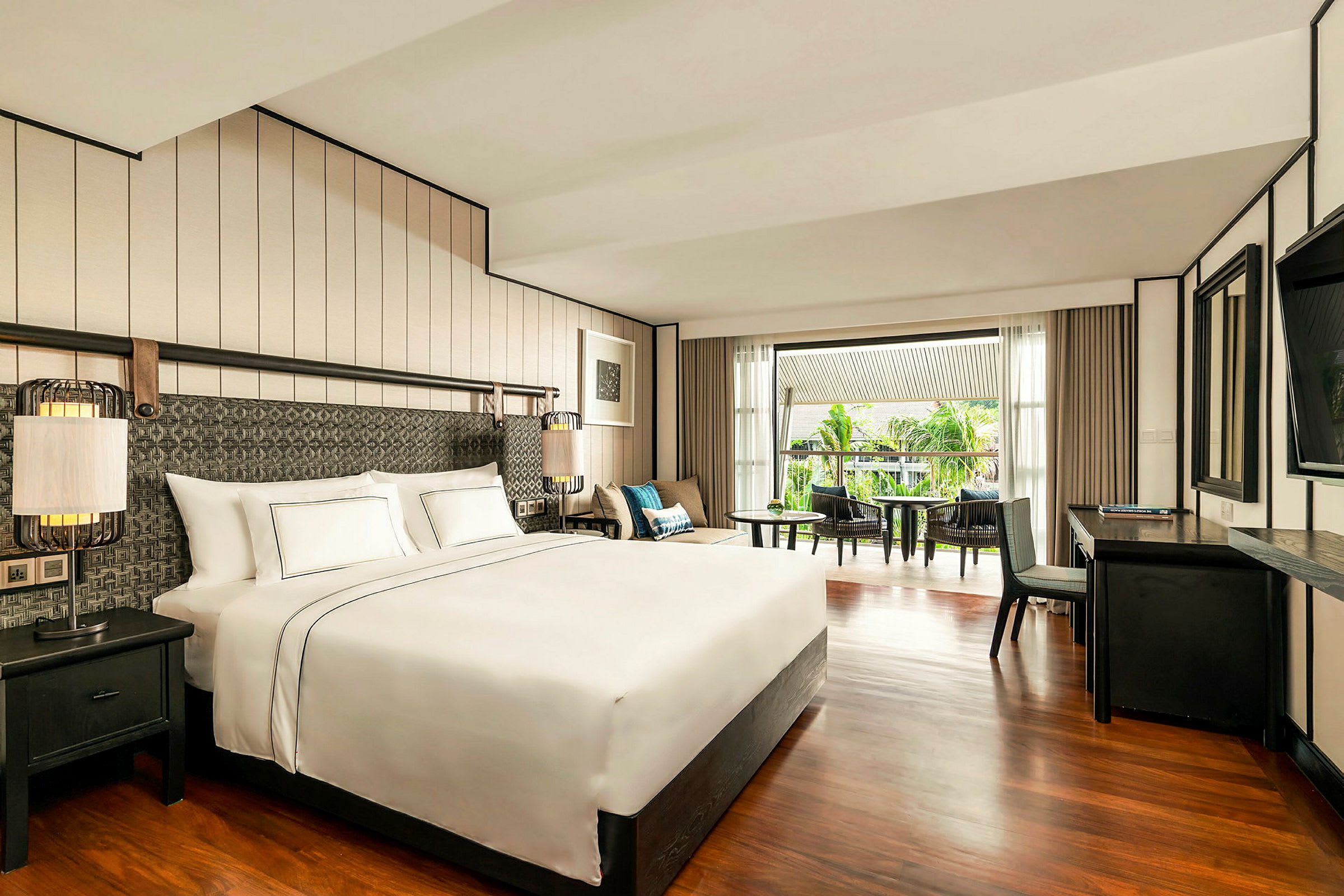 Ett hotellrum med en stor dubbelsäng, skrivbord, samt en sittgrupp med utsikt över en balkong och grönska utanför. (AI-genererad text)