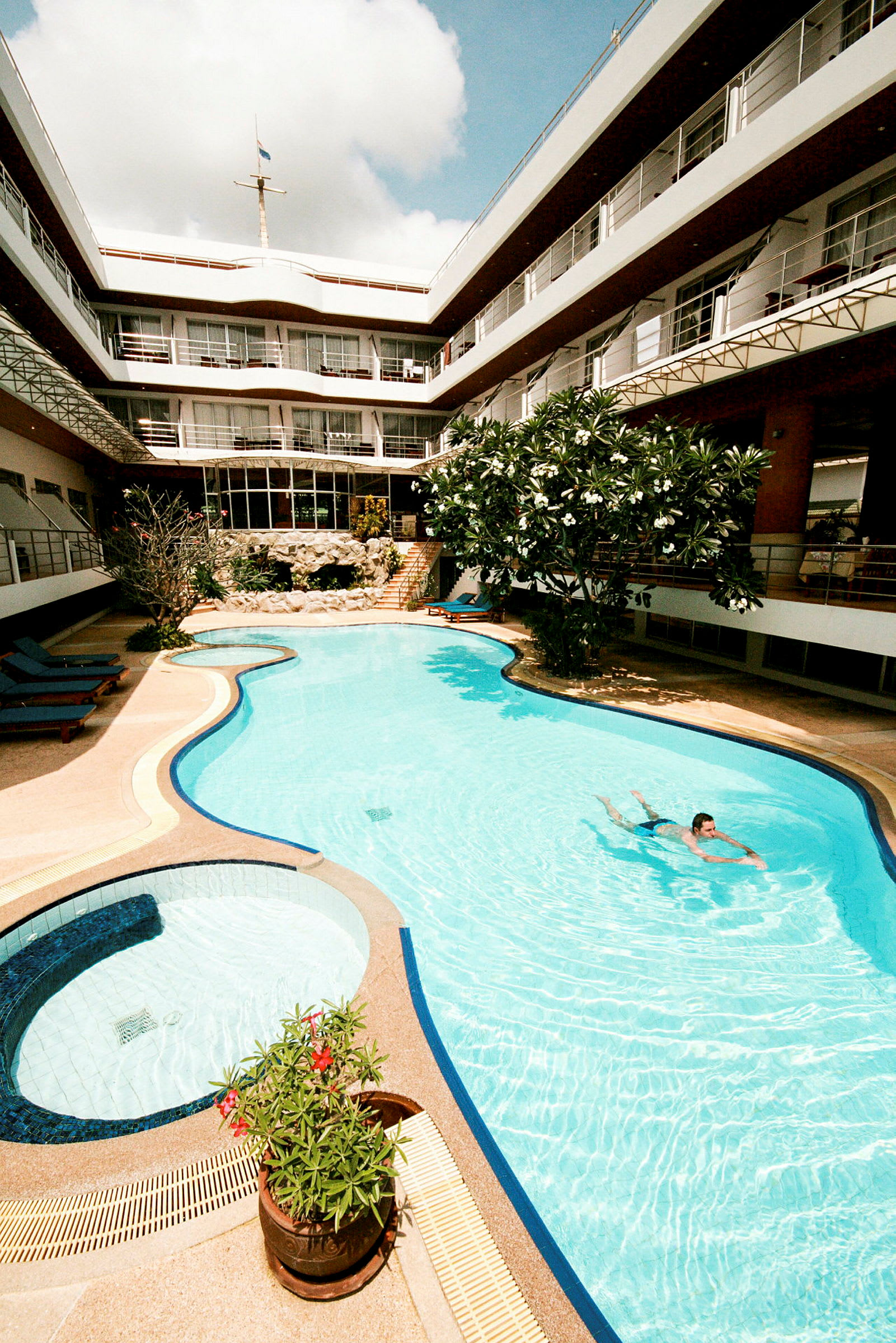 En pool omgiven av en flervåningsbyggnad med balkonger. Det finns solstolar och gröna växter runt poolen. (AI-genererad text)