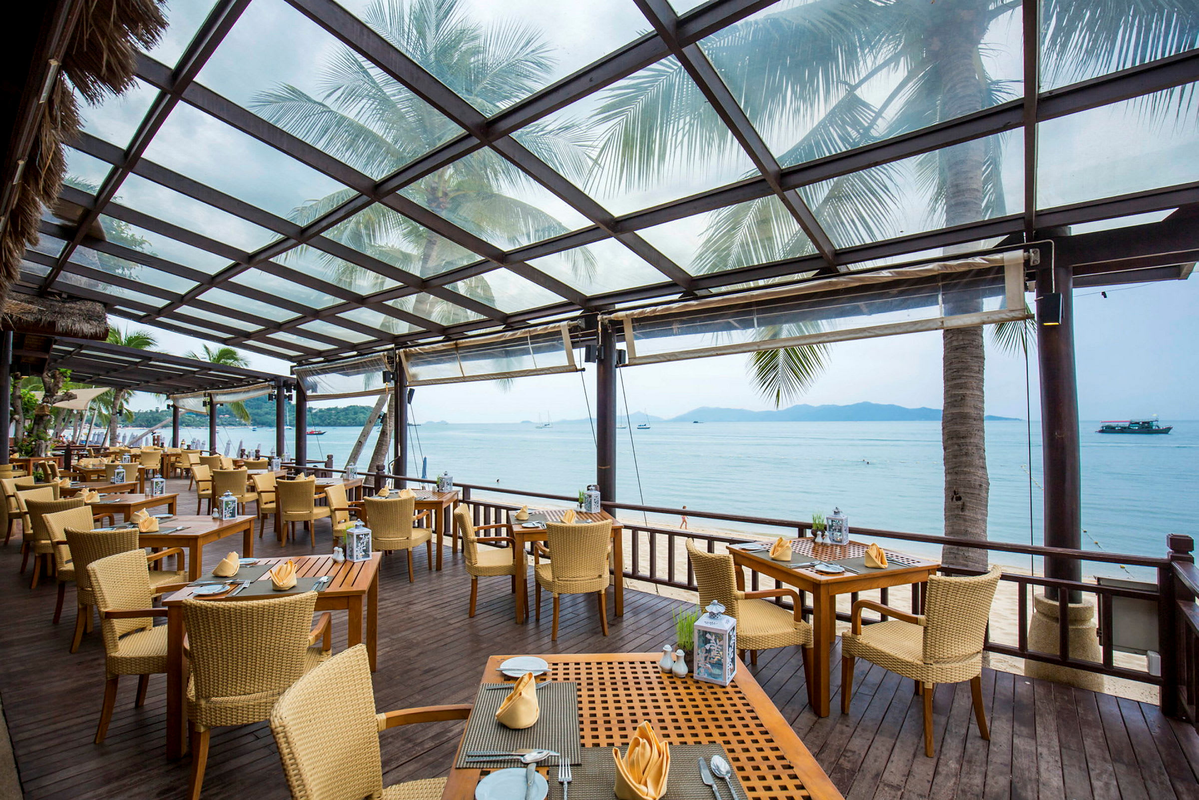 En restaurang med uteservering under ett tak av glas. Stolar och bord är uppställda på ett trädäck med en utsikt över stranden och havet. (AI-genererad text)