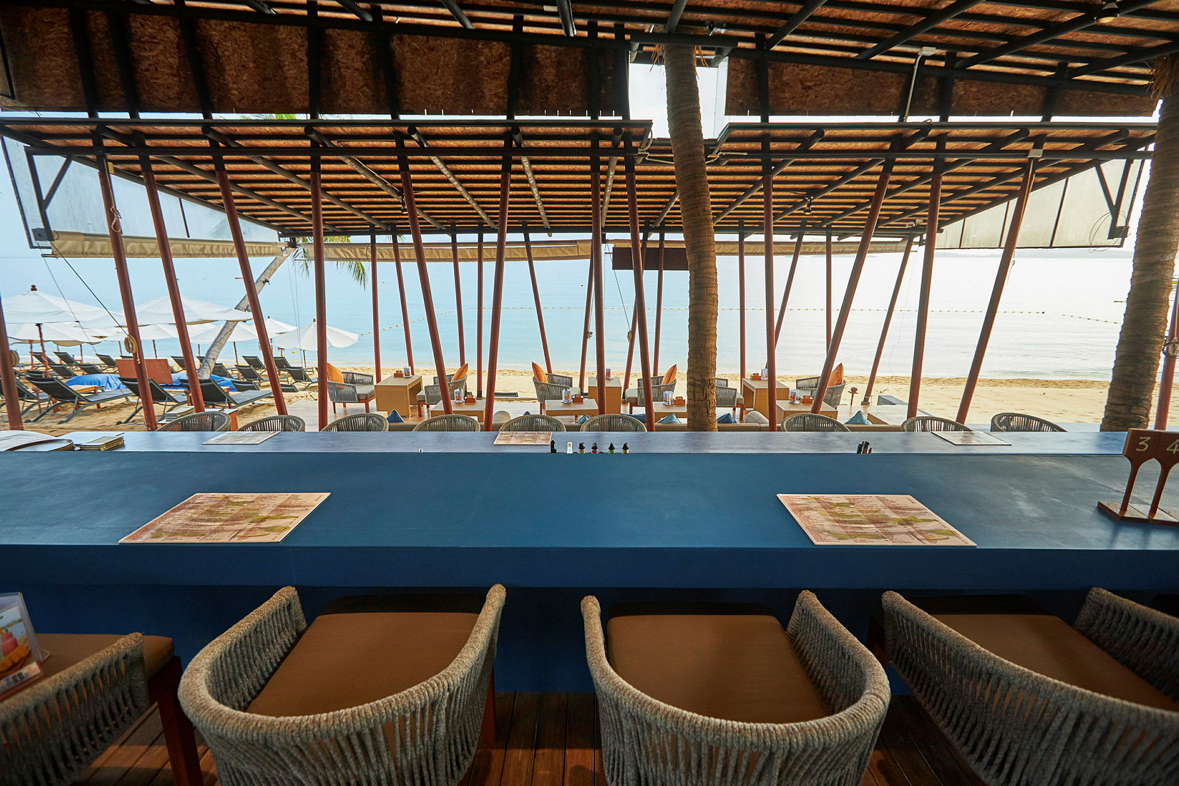 En restaurang med utsikt över stranden och havet, med bord och sittplatser under ett tak. (AI-genererad text)