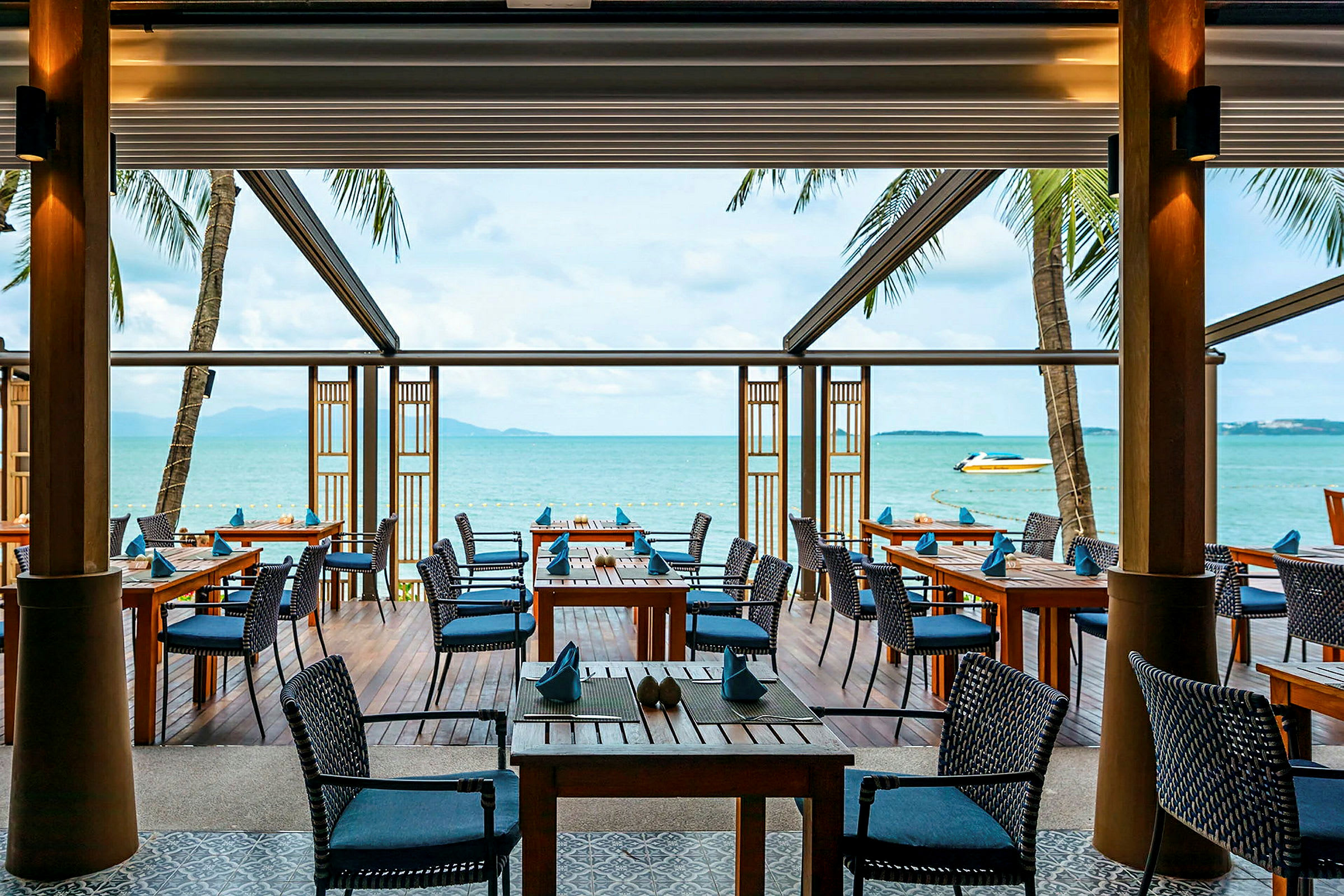 En utomhusrestaurang med träbord och stolar ligger vid havet och erbjuder en utsikt mot vattnet och en båt. (AI-genererad text)