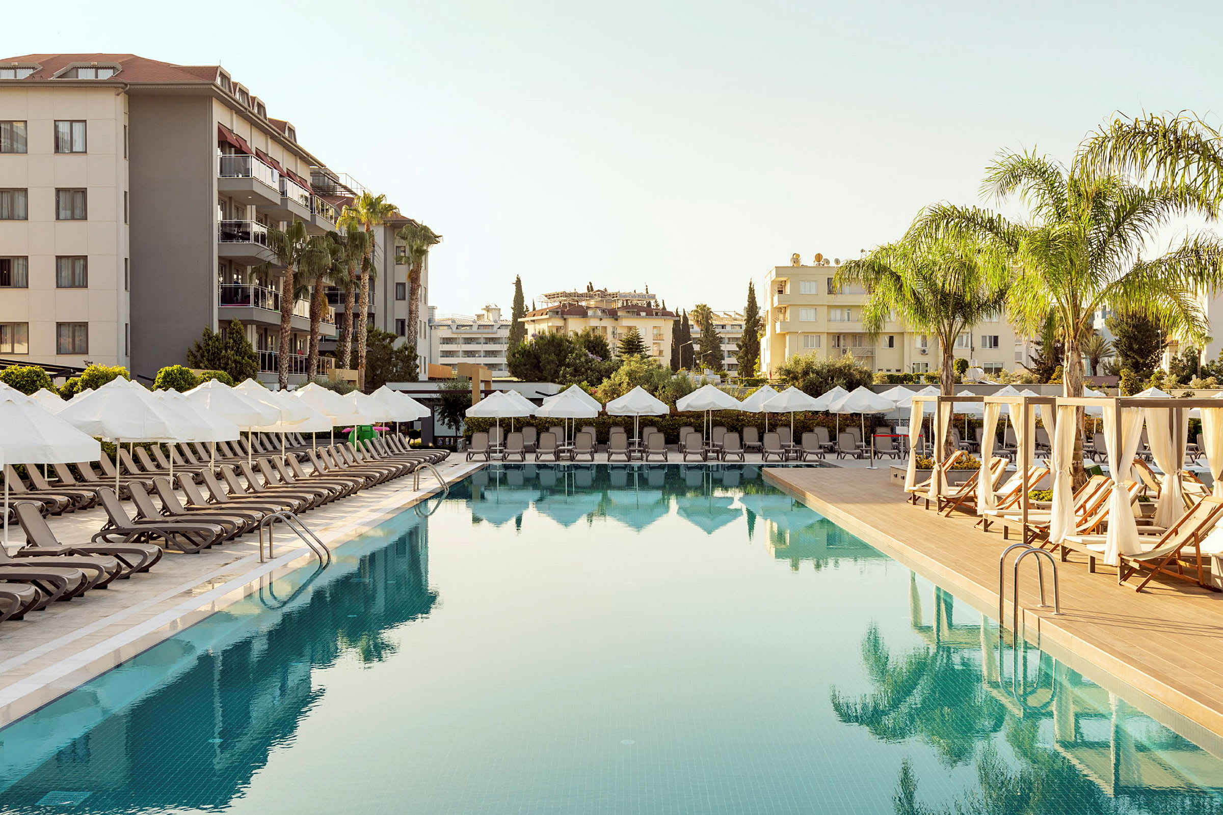 En hotellpool med solstolar och parasoller vid sidan, omgiven av byggnader och palmer. (AI-genererad text)