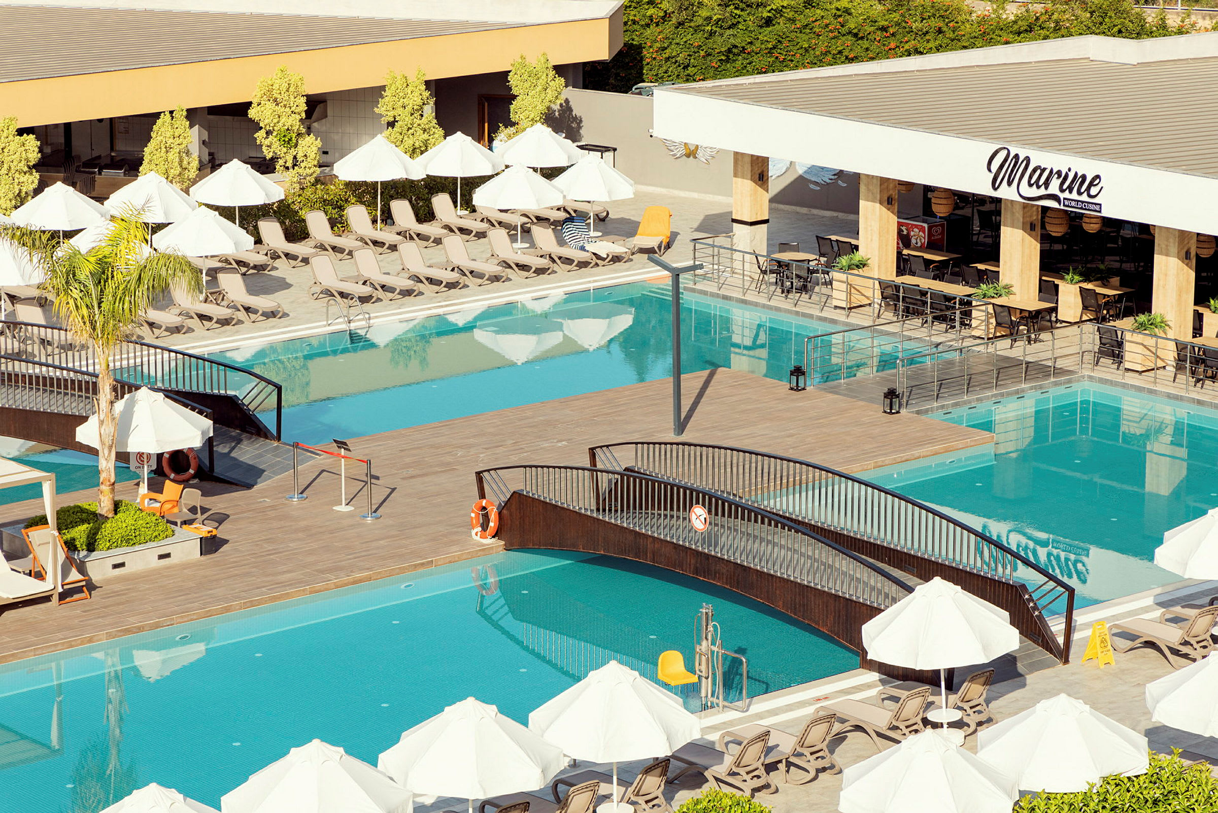 Poolområde med solstolar, parasoller, en gångbro över poolen och en restaurang med sittplatser. (AI-genererad text)