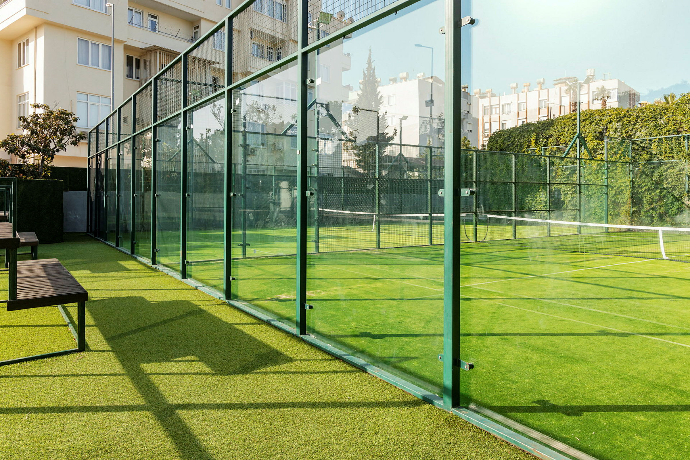 En utomhus padelbana omgiven av glasväggar, med grön konstgräsbeläggning och omgivande byggnader. (AI-genererad text)