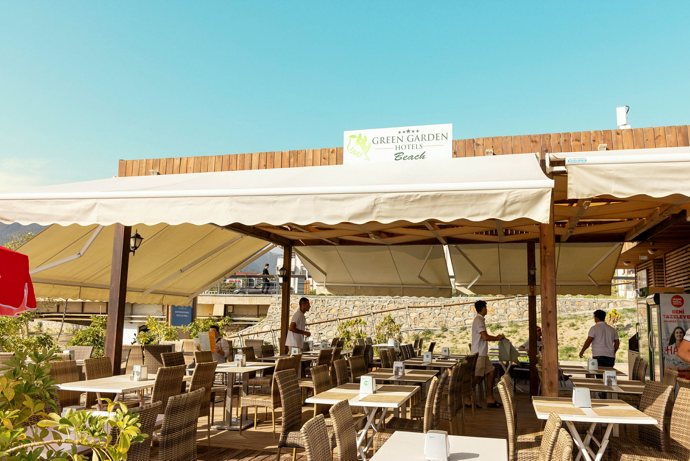 En uteservering med bord och stolar, med överliggande segeltak och en skylt med texten 'Green Garden Hotels Beach'. (AI-genererad text)