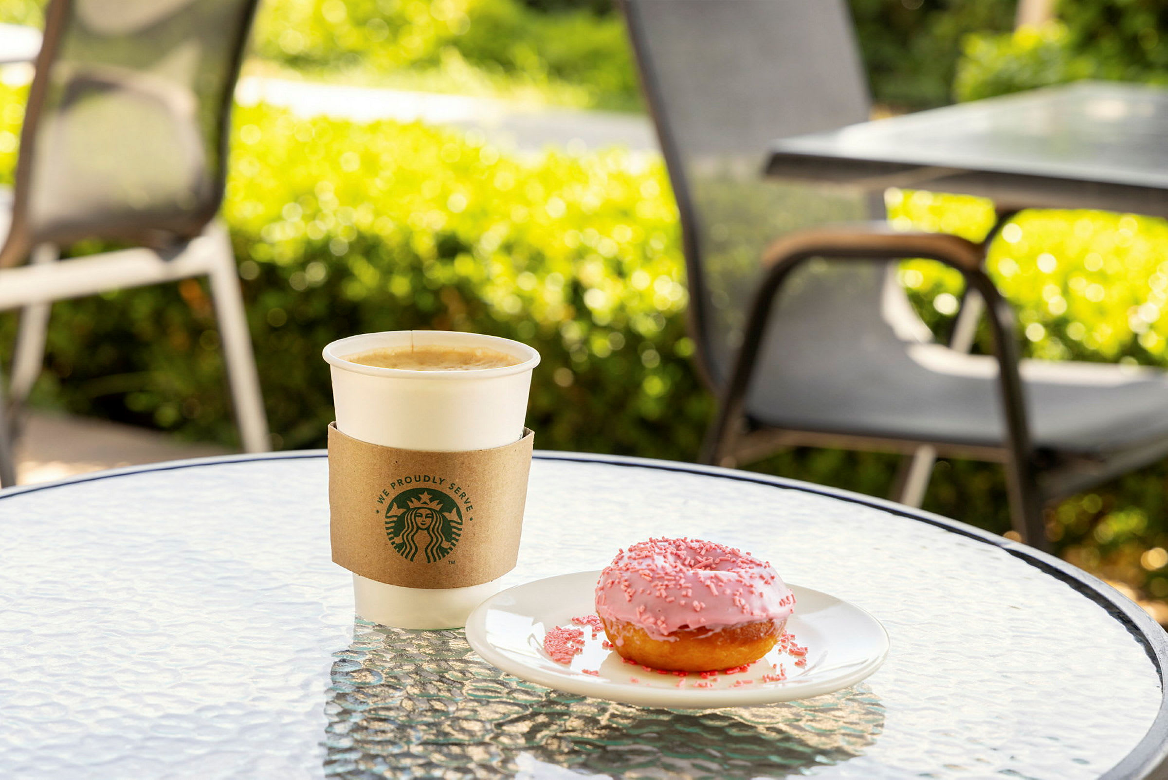 En kopp kaffe och en rosa munk på ett bord utomhus. (AI-genererad text)