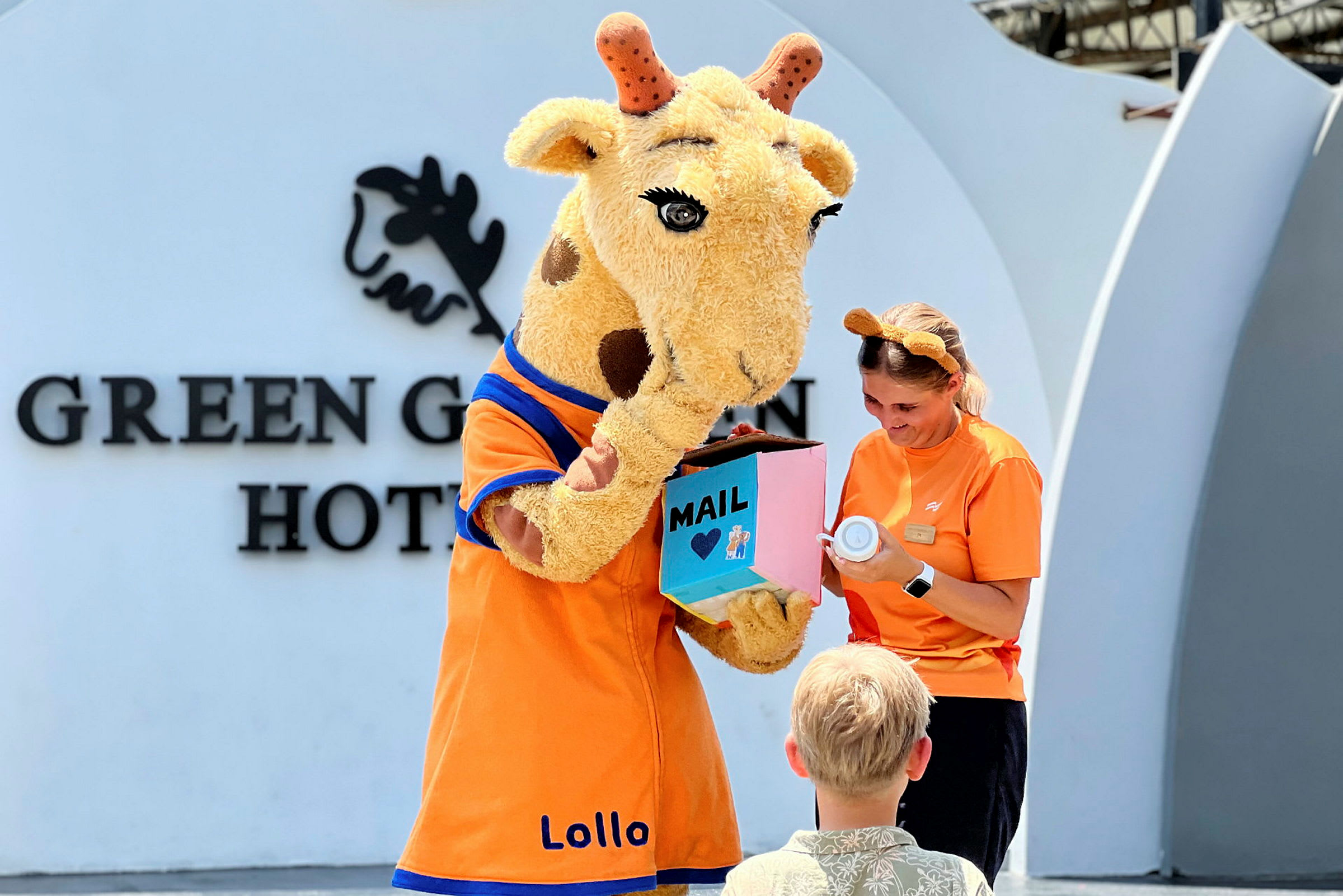 En person i en giraffkostym håller i en färgglad låda framför ett barn. (AI-genererad text)