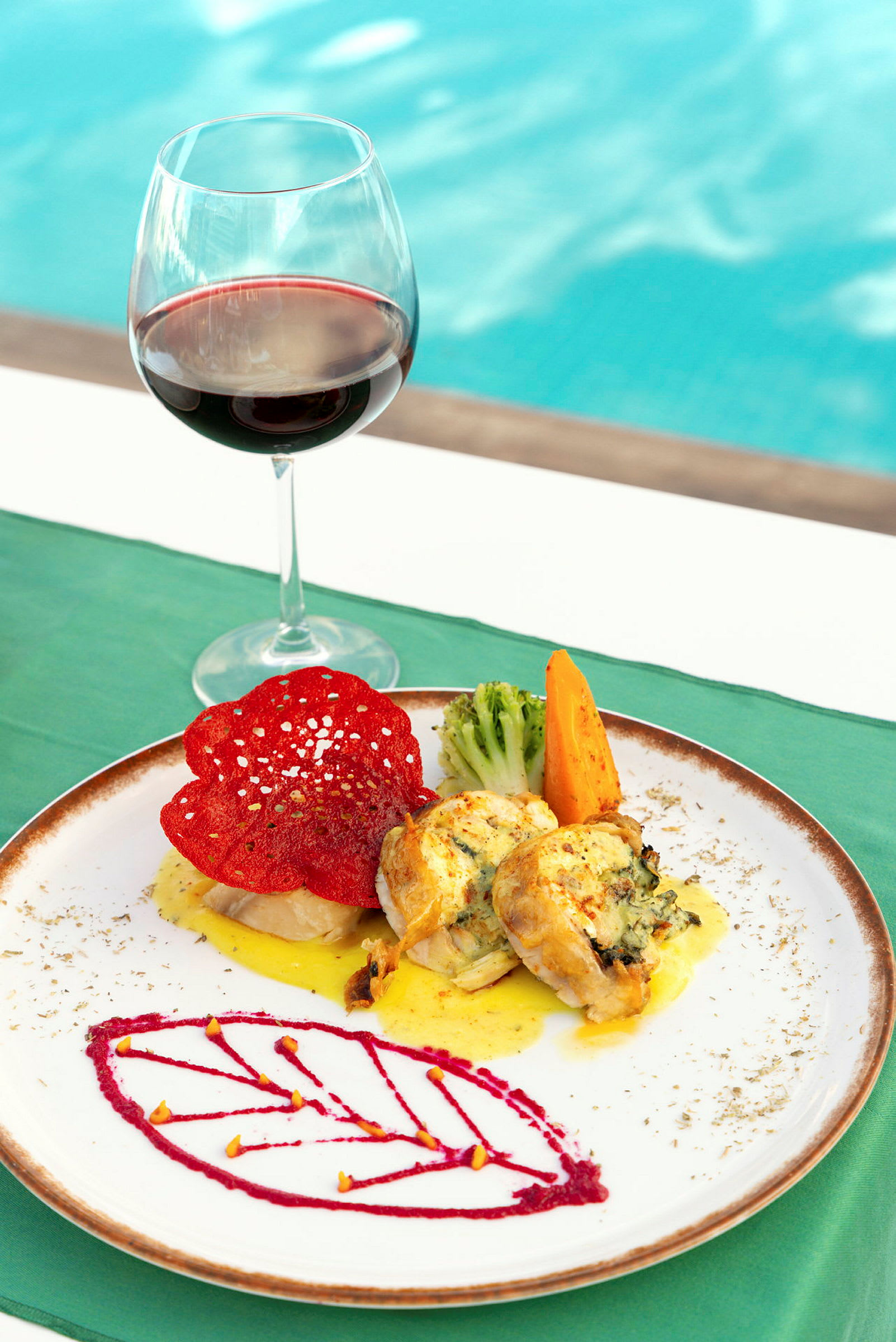En tallrik med en elegant måltid består av grönsaker och fylld kyckling bredvid ett glas rött vin, med en pool i bakgrunden. (AI-genererad text)