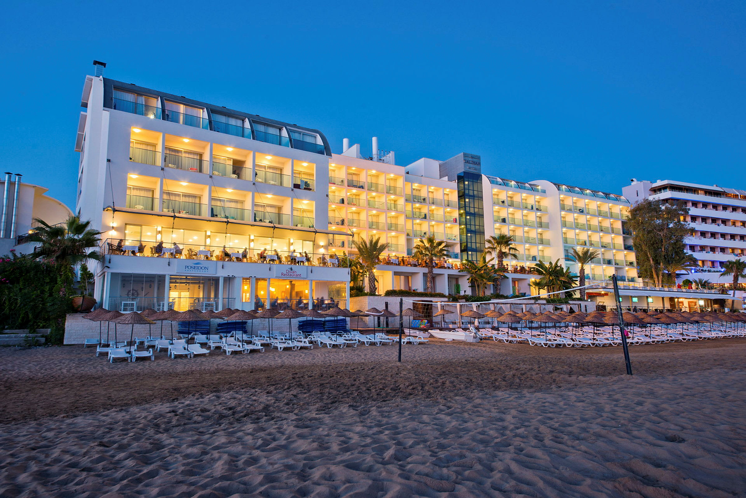 Hotellområde med upplyst fasad vid stranden, solstolar och parasoller i förgrunden. (AI-genererad text)