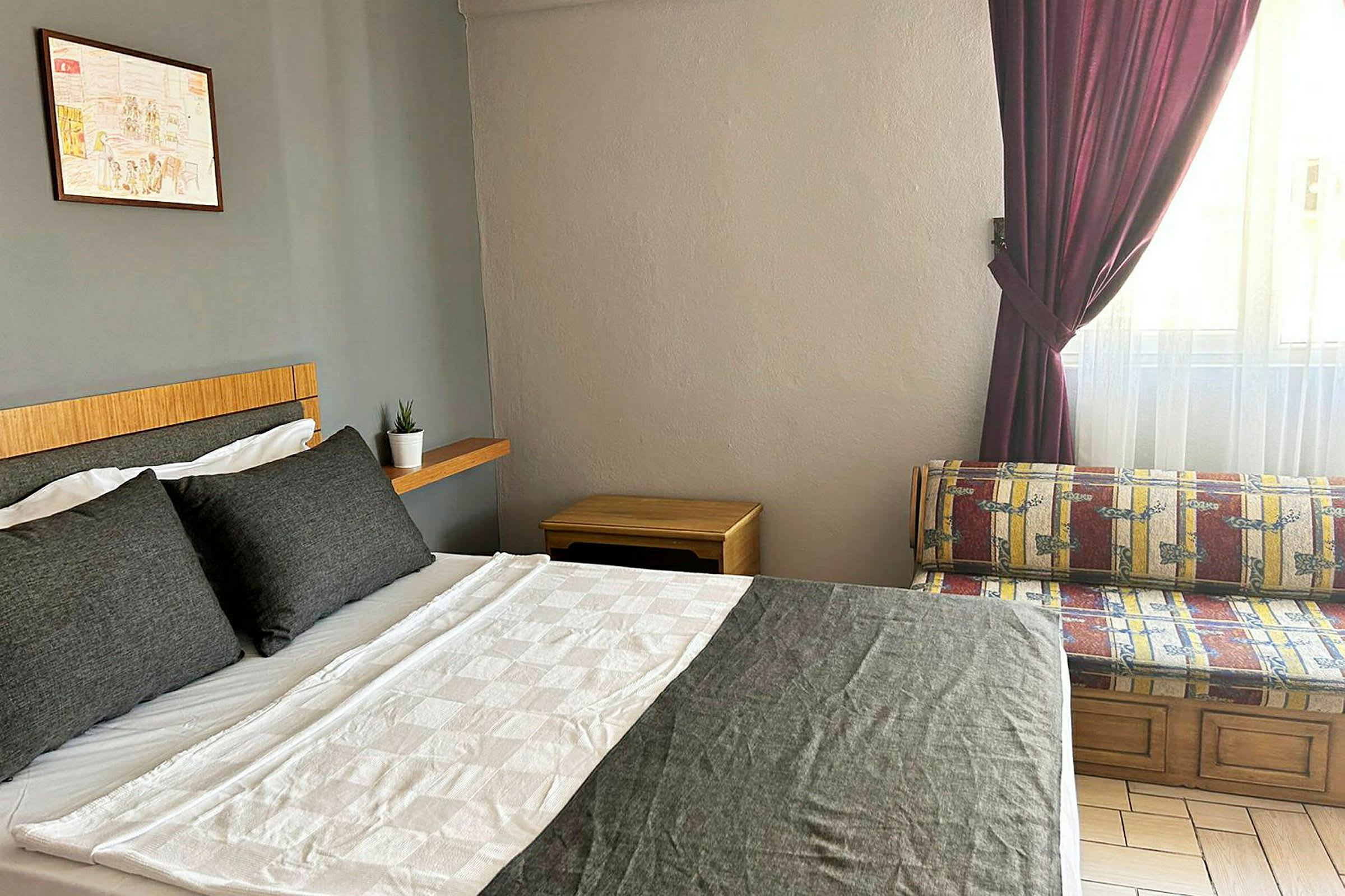 Ett sovrum med en dubbelsäng med grå och vita sängkläder. Bredvid sängen finns en hylla med en liten krukväxt. En fönstersoffa är placerad nära ett fönster med mörklila gardiner. (AI-genererad text)
