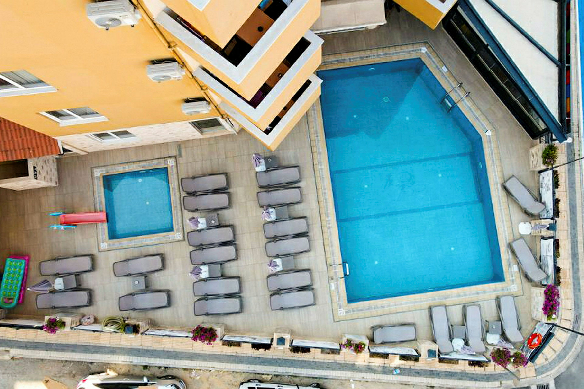 Bild av en utomhuspool och en mindre barnpool, omgiven av solstolar och parasoller vid ett hotell. Högerkant syns en gata. (AI-genererad text)