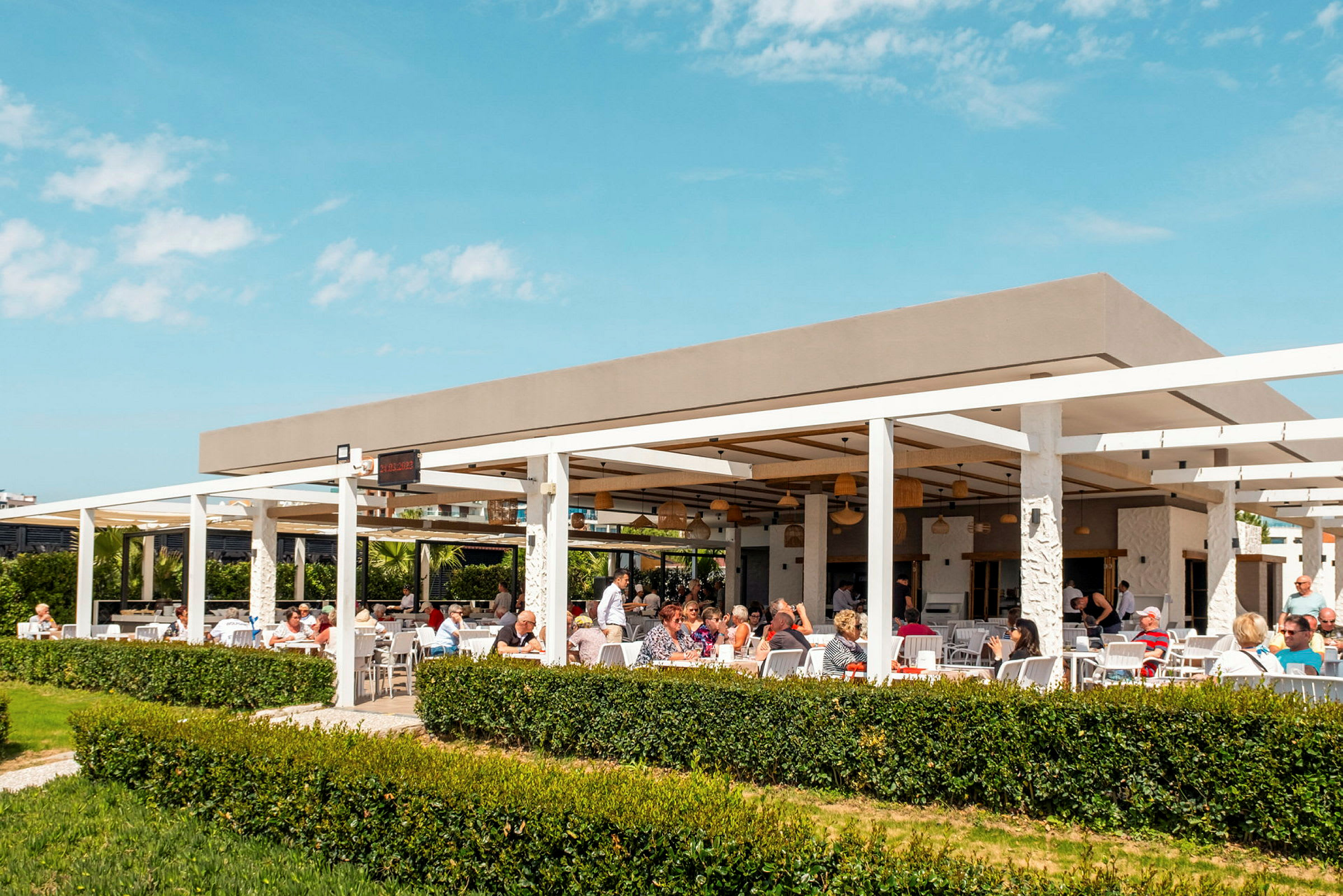 Utomhusterrass med sittplatser och människor som äter under en pergola. (AI-genererad text)