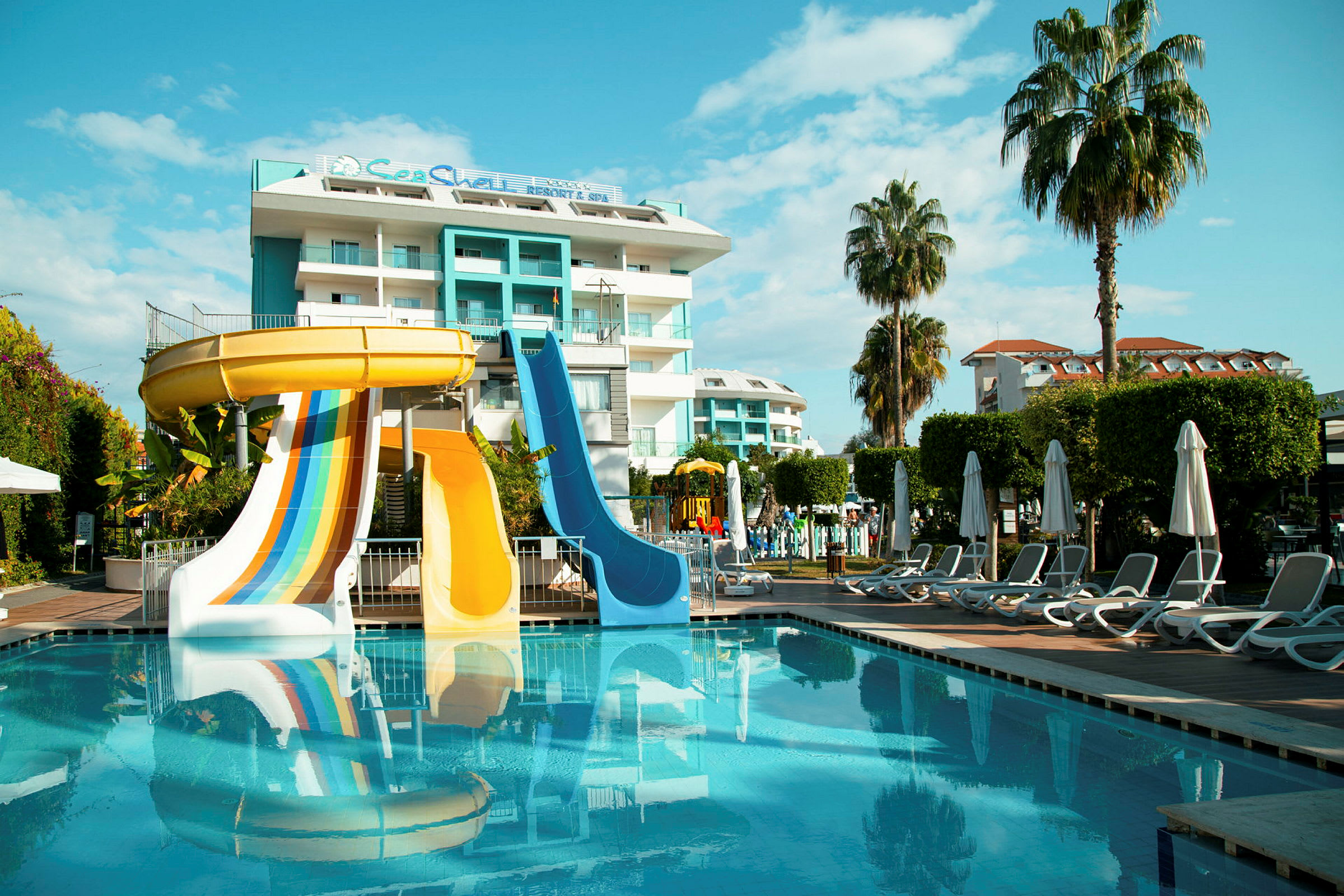 En hotellbyggnad med pool och vattenrutschbanor framför, omgivet av solstolar och palmer. (AI-genererad text)