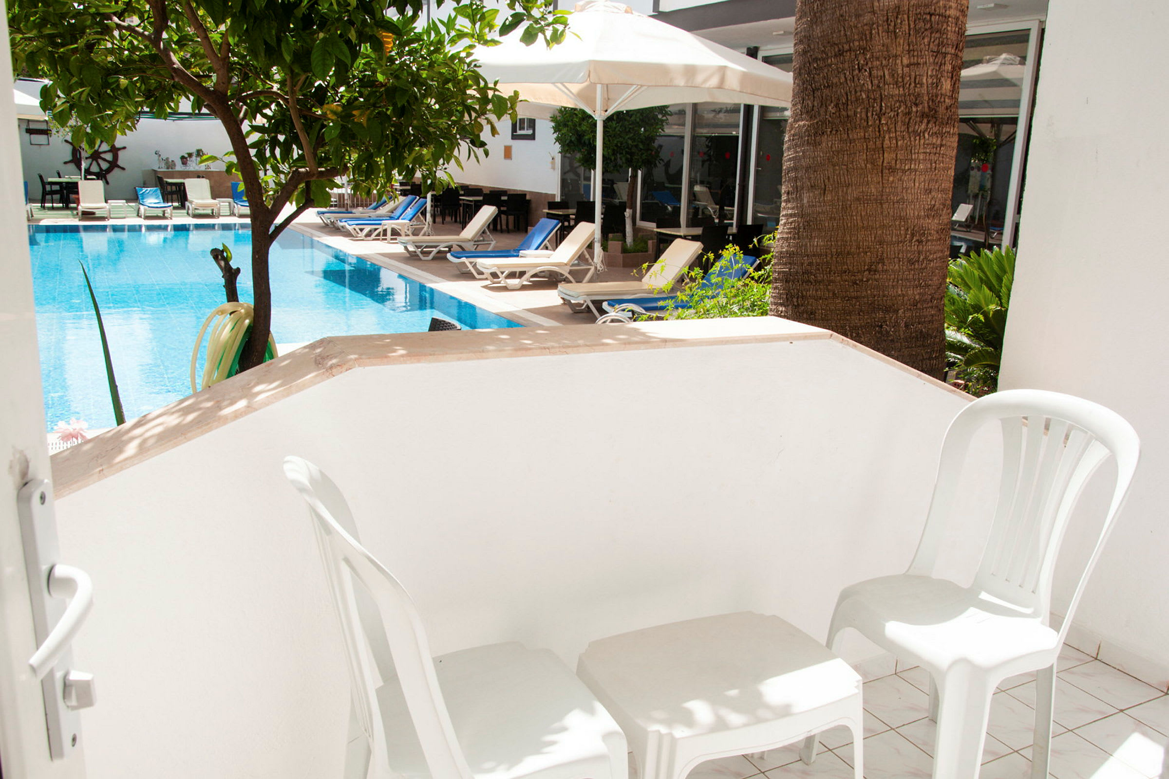 En balkong med två vita plaststolar och ett bord, med utsikt över en pool och solstolar. (AI-genererad text)