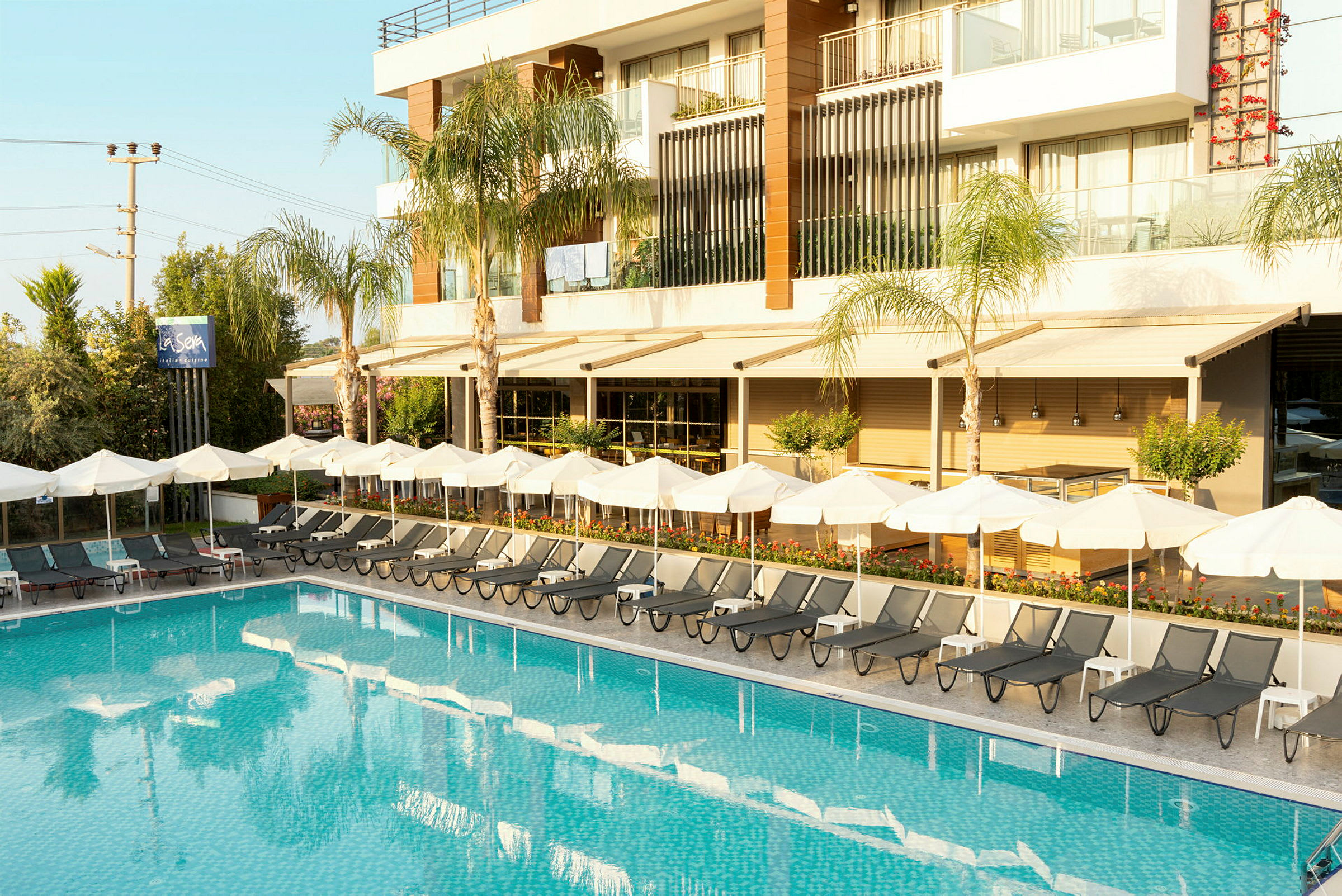 Utomhusområde med en pool, solstolar och parasoller framför en modern byggnad. (AI-genererad text)
