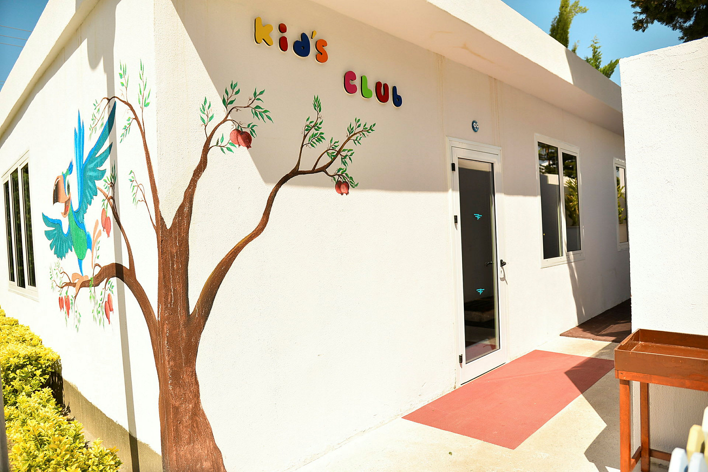 Vit byggnad med dekorationer av ett målat träd och en färgglad papegoja på fasaden, med texten 'Kid's Club' ovanför dörren. (AI-genererad text)