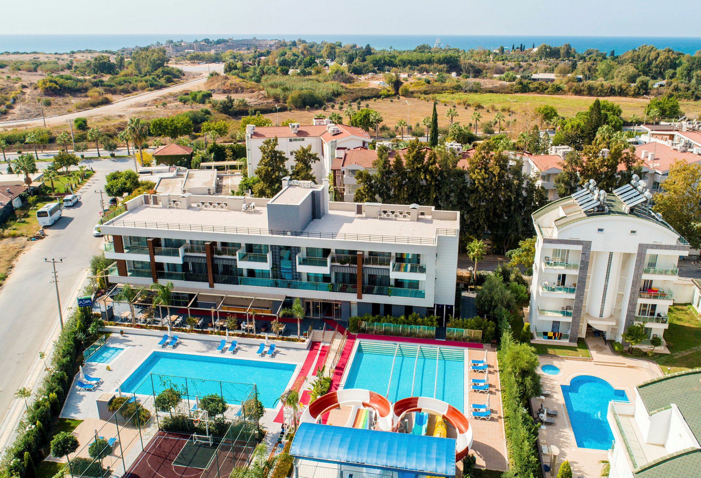 Ett hotellkomplex med pooler, vattenrutschbanor och en idrottsplan. Havet och landskap i bakgrunden. (AI-genererad text)