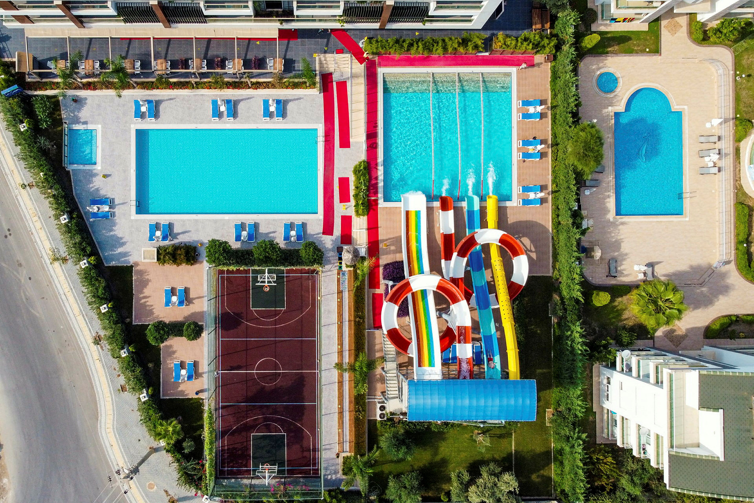 En fågelperspektivbild på ett hotellområde med flera pooler, solstolar, vattenrutschbanor och en basketplan. (AI-genererad text)