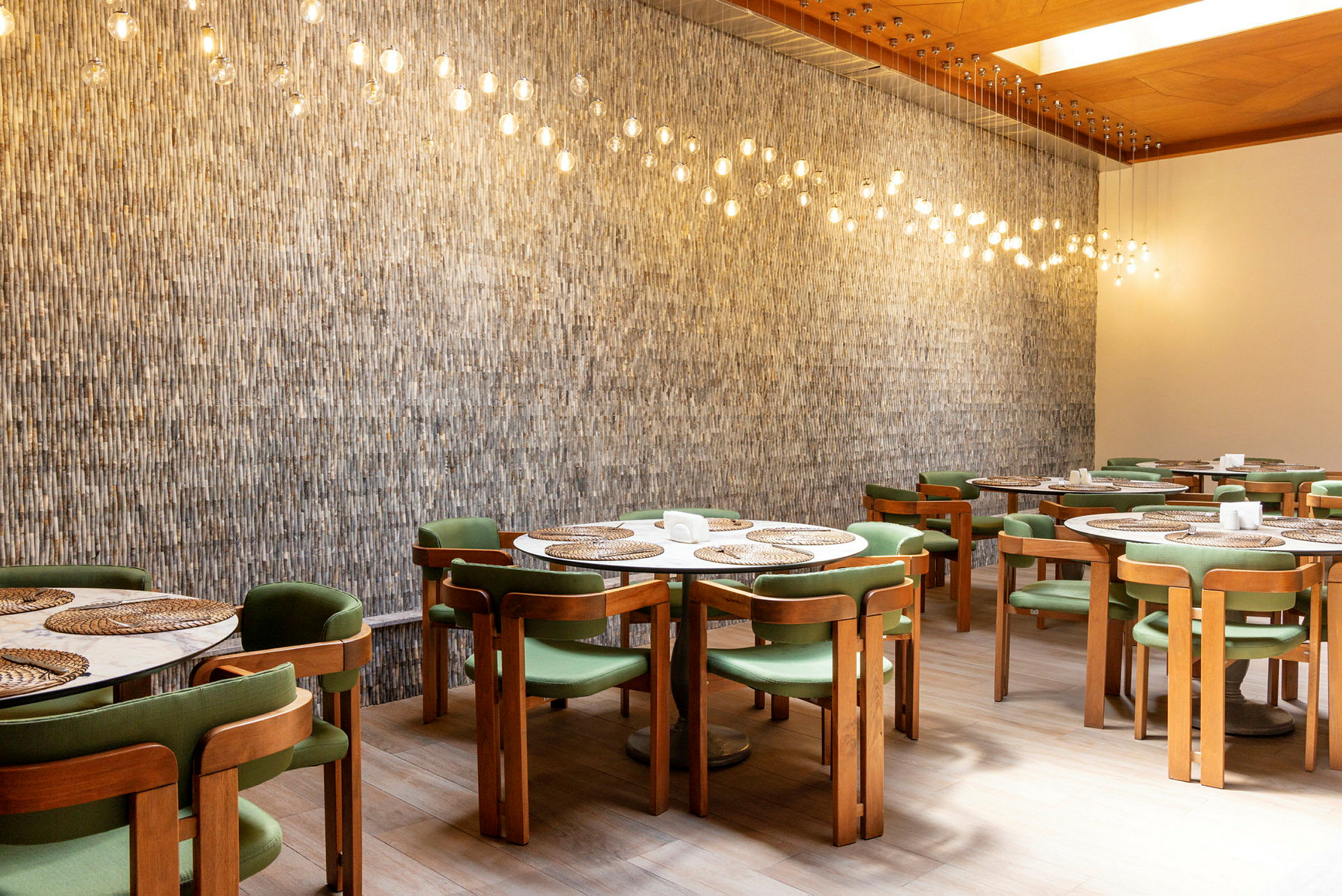 En inomhusrestaurang med trästolar och gröna sitsar, runt bord med vassmattor och hängande lampor med glödlampor ovanför. (AI-genererad text)