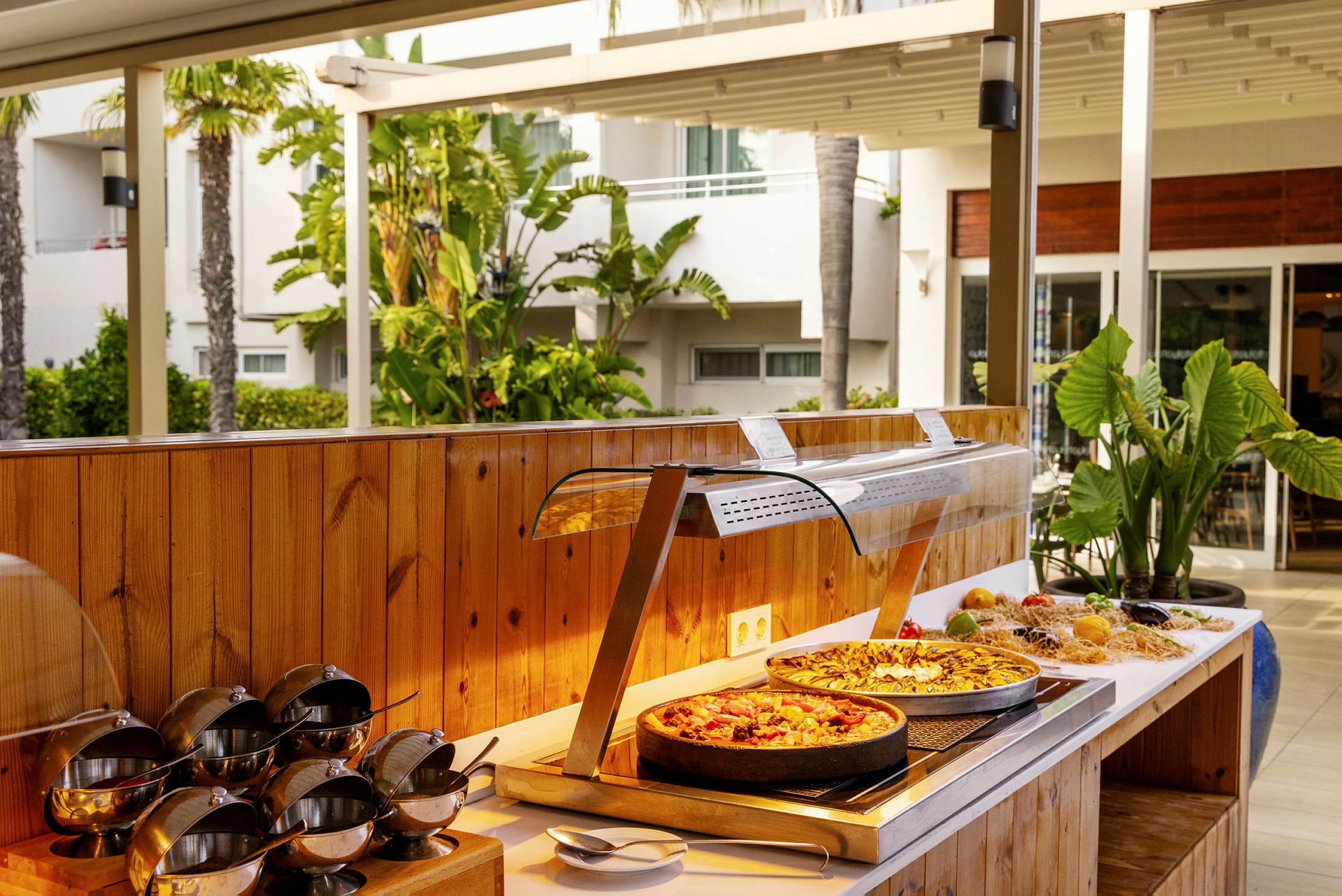 En buffé med mat, inklusive två pizzor, omgiven av gröna växter och en utomhusmiljö. (AI-genererad text)