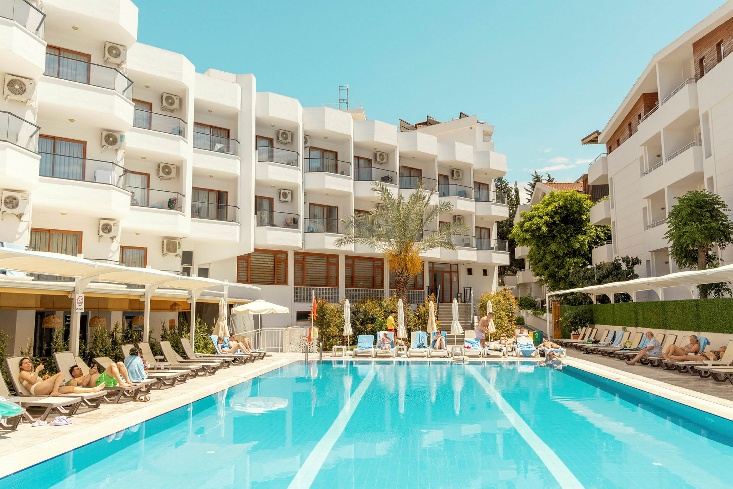 Ett hotell med vit fasad och balkonger syns i bakgrunden. Framför byggnaden ligger en utomhuspool omgiven av solsängar och parasoller, där människor kopplar av. (AI-genererad text)