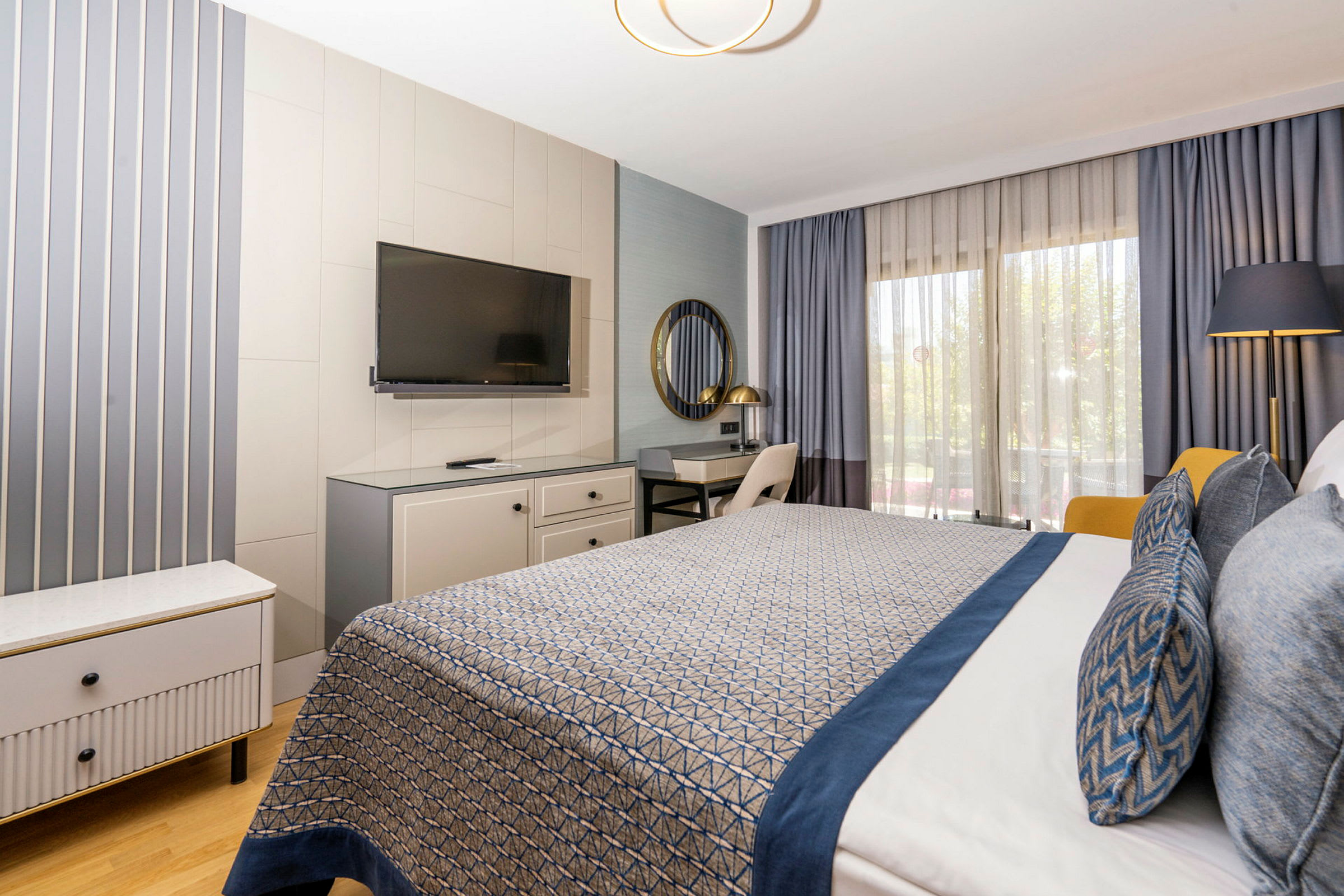 Hotellrum med modernt inredd interiör, inklusive säng med blå detaljer, skrivbord, TV och stora fönster med gardiner. (AI-genererad text)