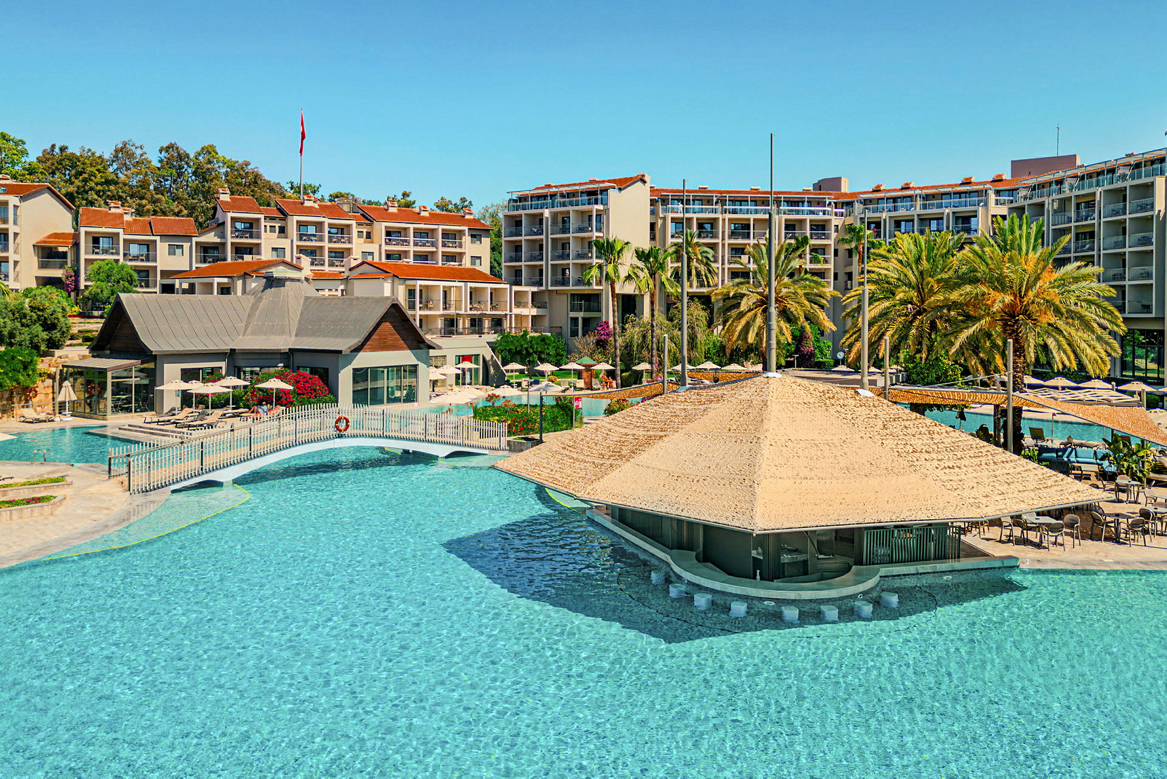 Bild av en hotellresort med en stor swimmingpool, övertäckt poolbar och omgivande byggnader samt palmer. (AI-genererad text)