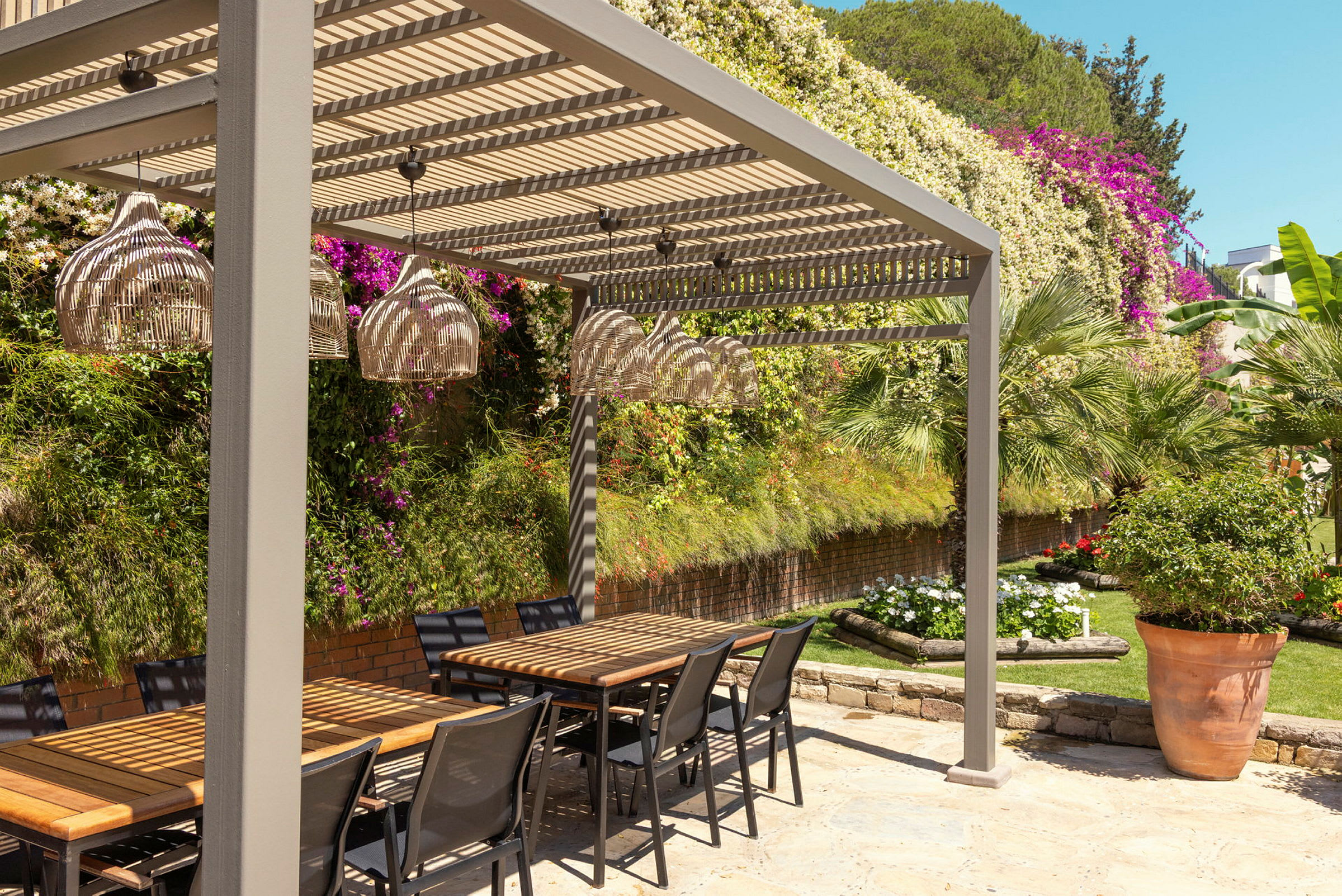 En sittgrupp utomhus med bord och stolar under en pergola, omgiven av grönska och blommor. (AI-genererad text)