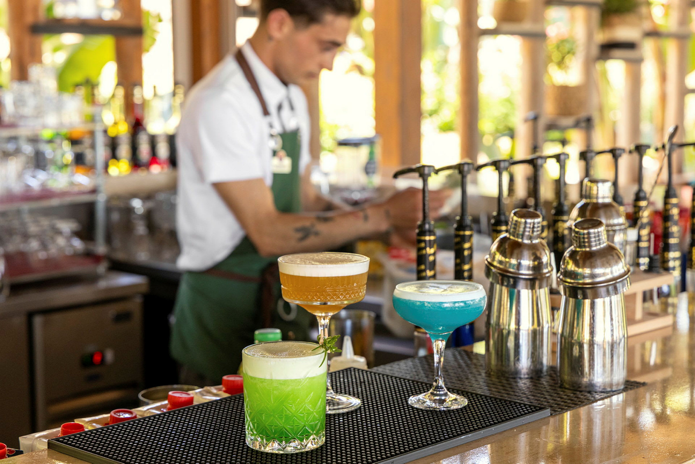 En bar med tre färgglada cocktails, shakers och en bartender i bakgrunden. (AI-genererad text)