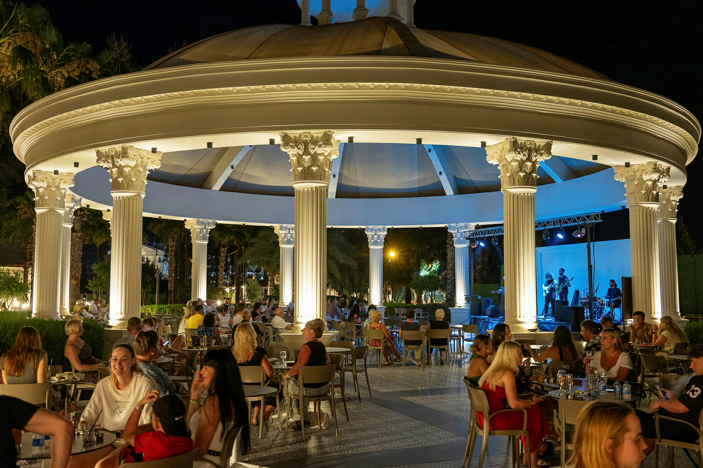 Utomhusrestaurang med konsert på kvällstid, med belysning och människor vid bord. (AI-genererad text)