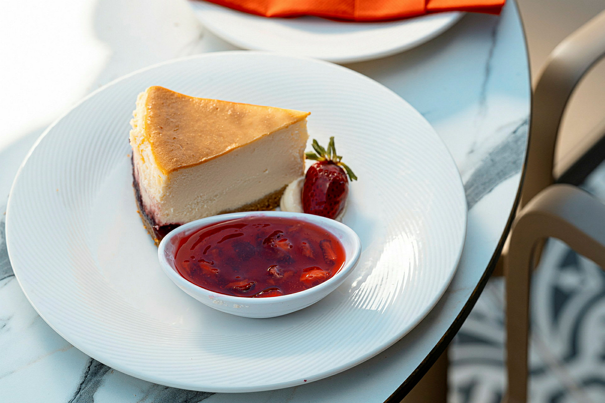 En portion cheesecake med tillhörande jordgubbssylt och en jordgubbe, serverad på en vit tallrik. (AI-genererad text)