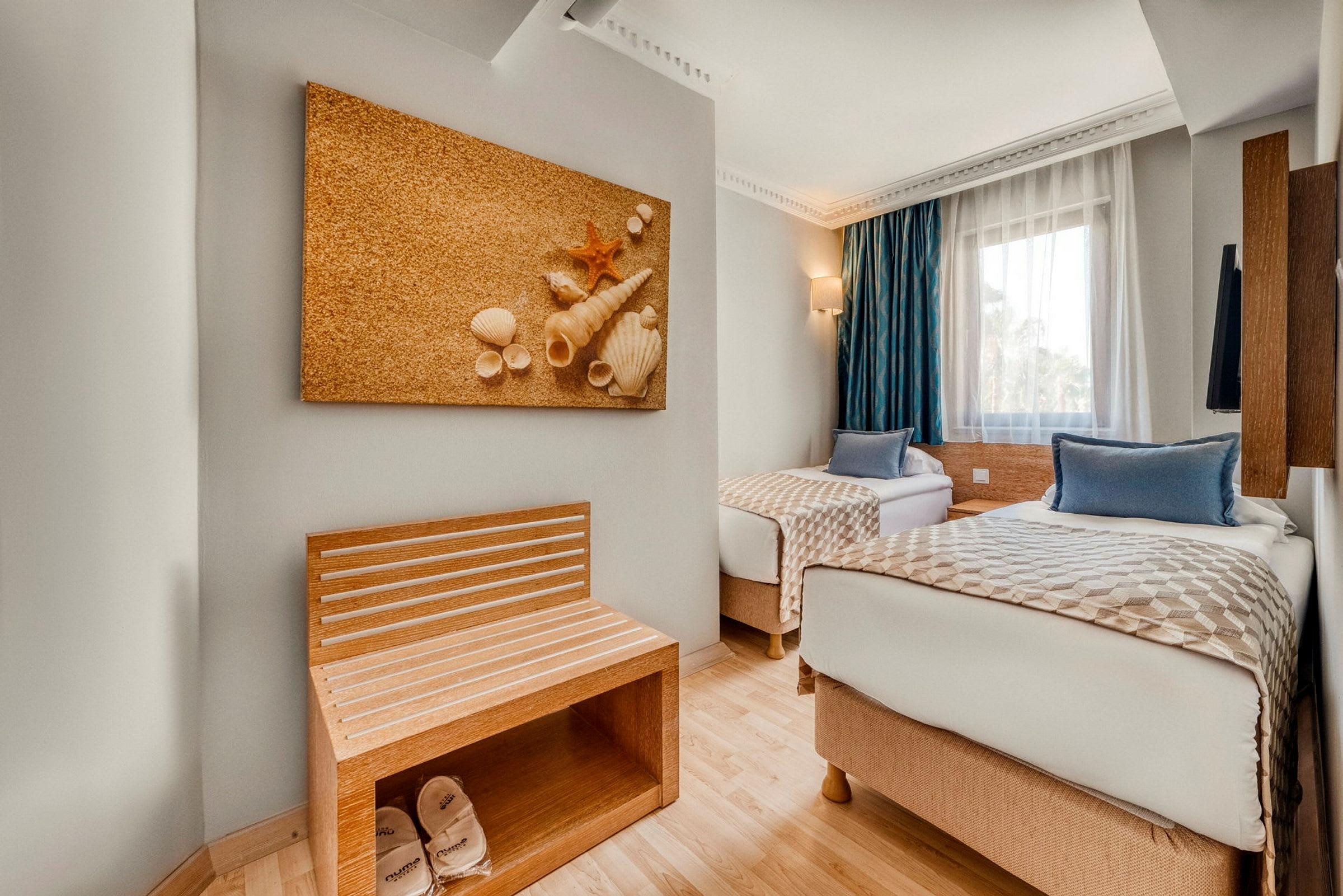 Två enkelbäddar i ett hotellrum med en väggdekoration av snäckskal och en sittbänk med tofflor. Bredvid finns ett fönster med blå gardiner. (AI-genererad text)
