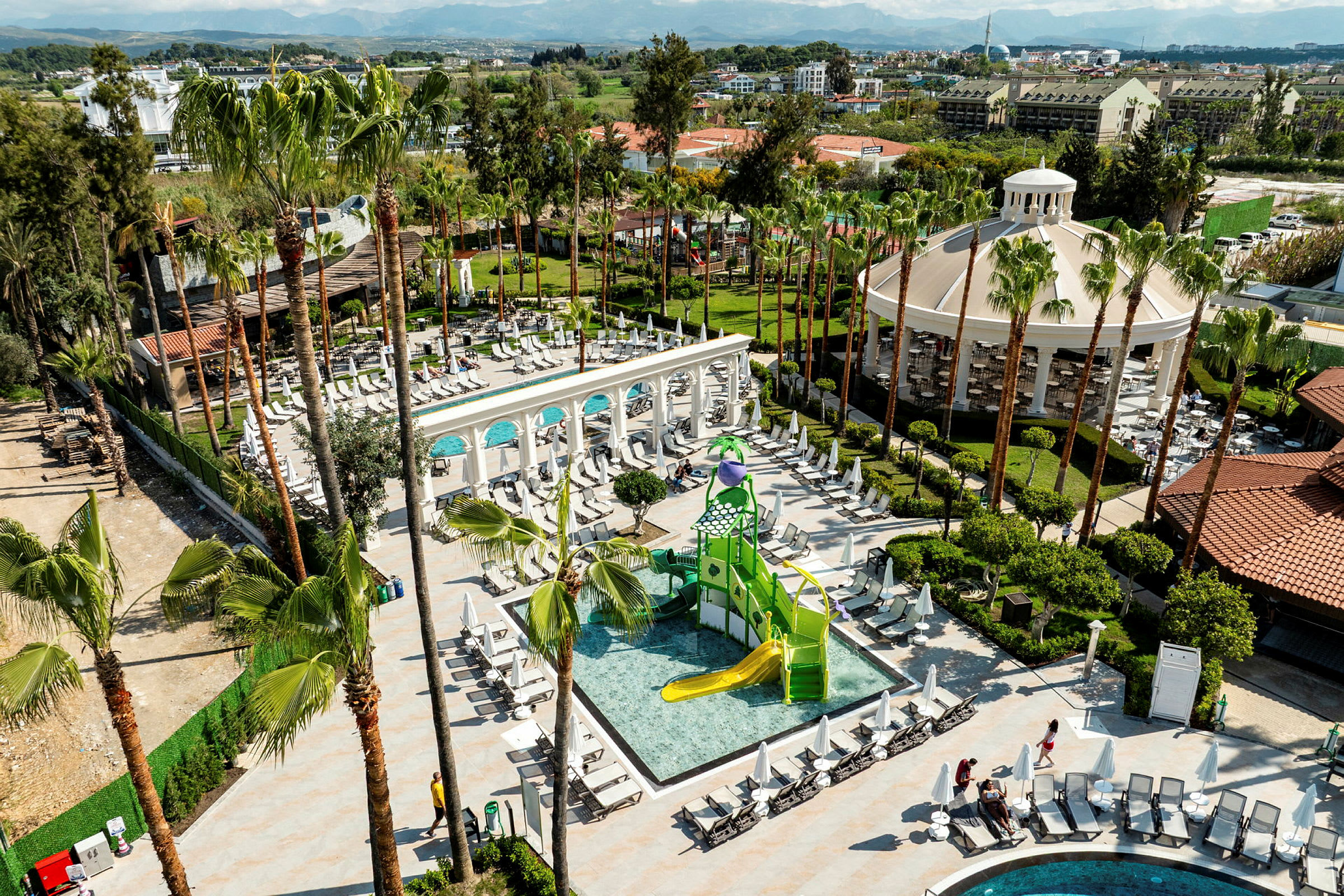 Bild av en resort med en barnpool, lekplats och omkringliggande solstolar samt grönska. (AI-genererad text)