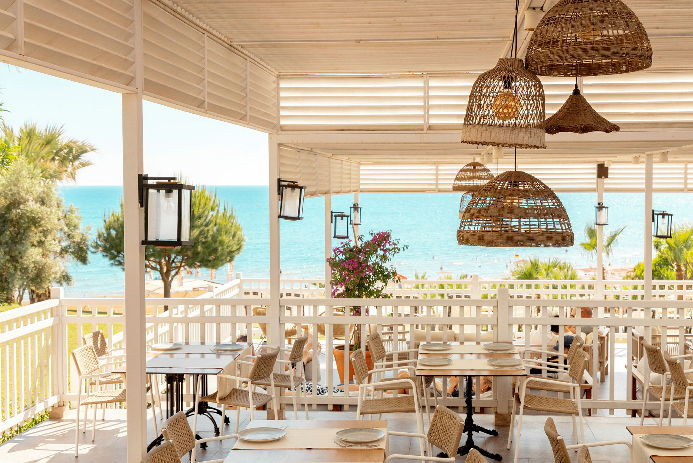 En strandnära restaurang med träbord, rottingstolar och utsikt över havet. (AI-genererad text)