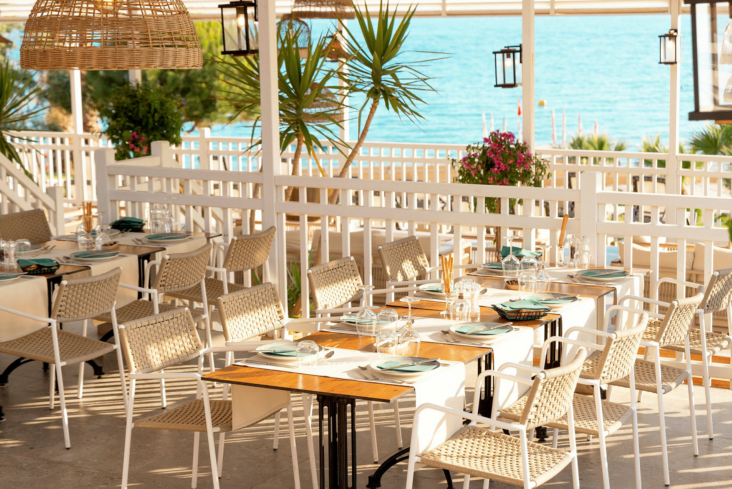 En utomhusrestaurang med bord uppdukade med tallrikar och glas. Utsikt över havet i bakgrunden. (AI-genererad text)