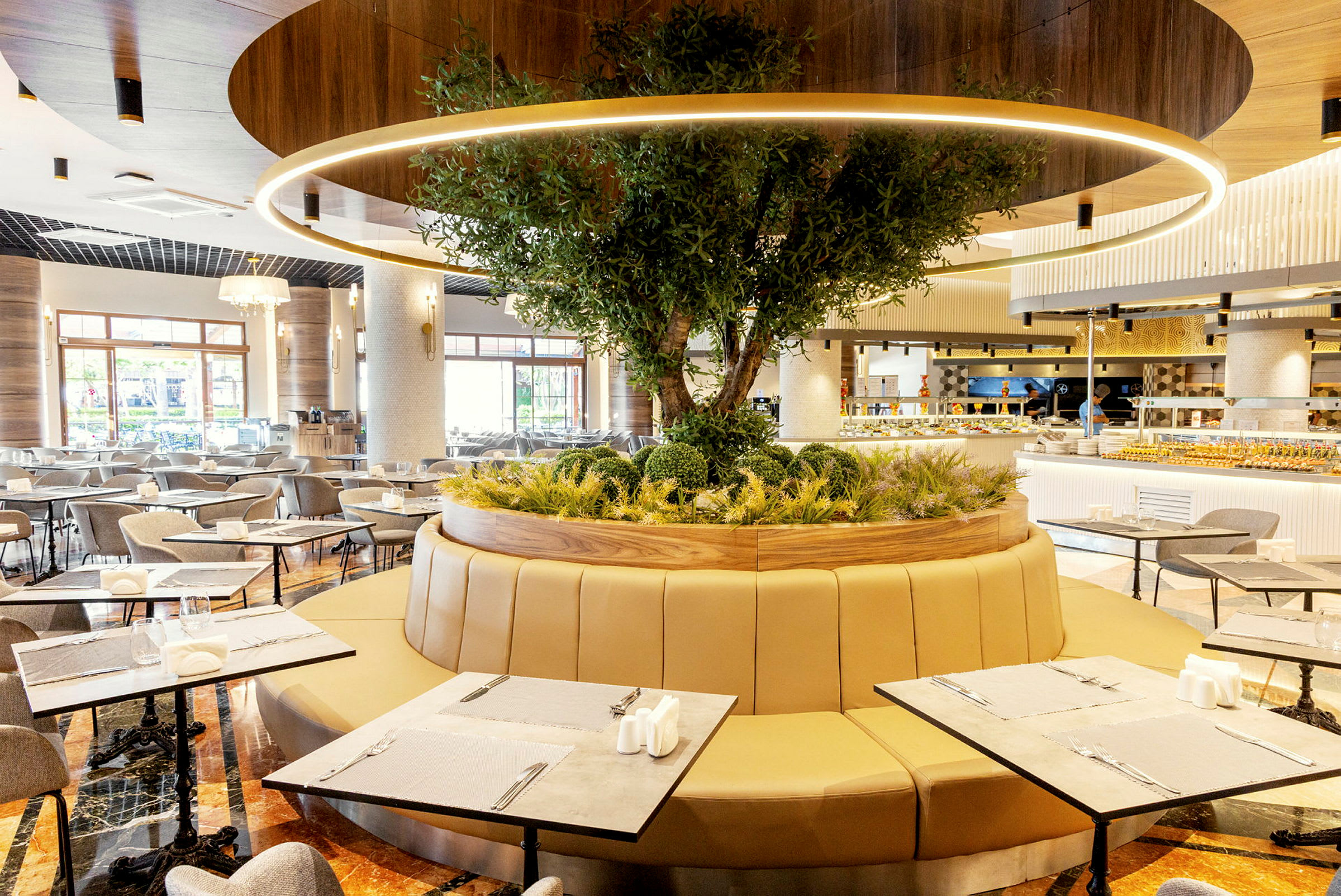 Ett modernt restaurangområde med sittplatser runt ett cirkulärt planteringsområde med ett träd. (AI-genererad text)