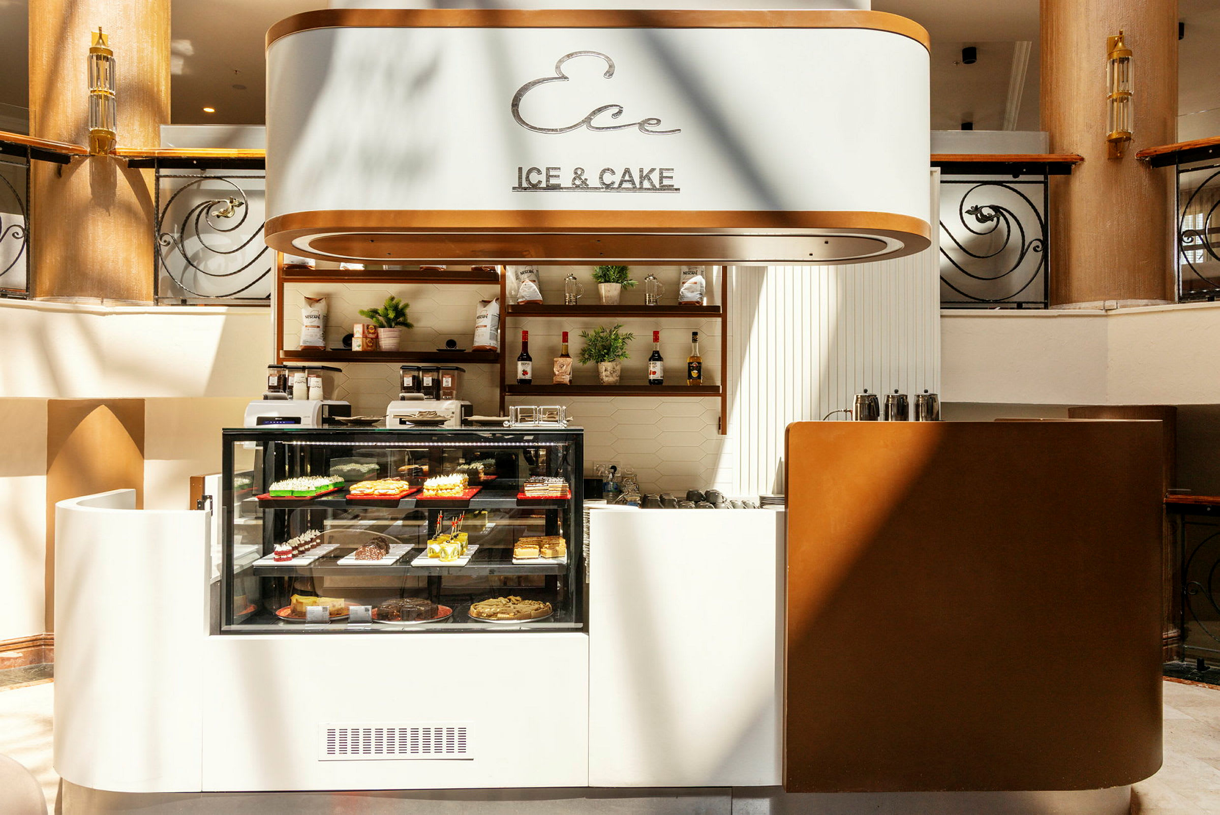 Ett kafé med glasmonter som visar olika bakverk, skyltar med texten 'Ice & Cake' och inredning i ljusa toner. (AI-genererad text)