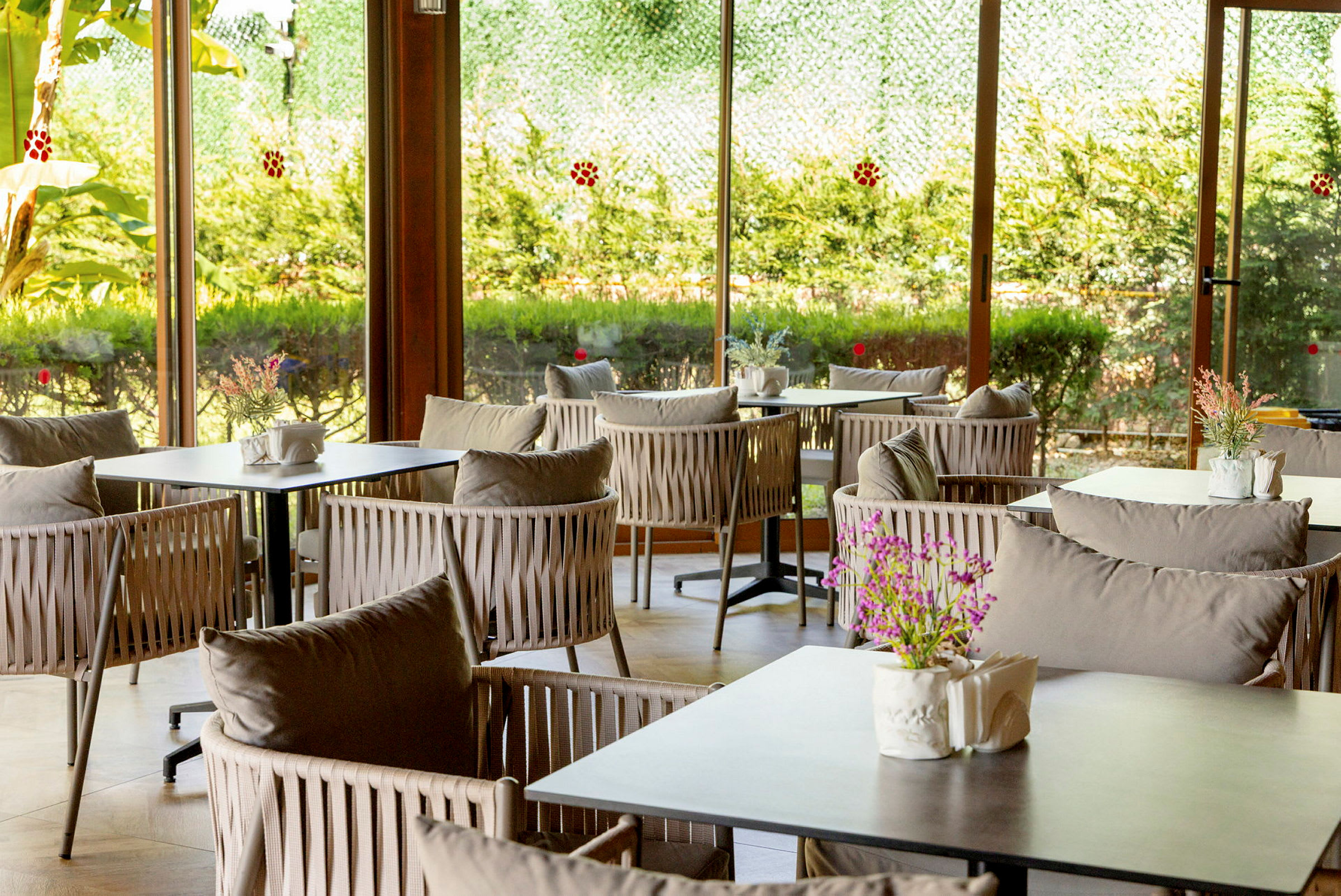 Ett luftigt och ljust café med moderna stolar runt bord, dekorerat med blommor och servettställ, beläget nära stora fönster som släpper in dagsljus. (AI-genererad text)