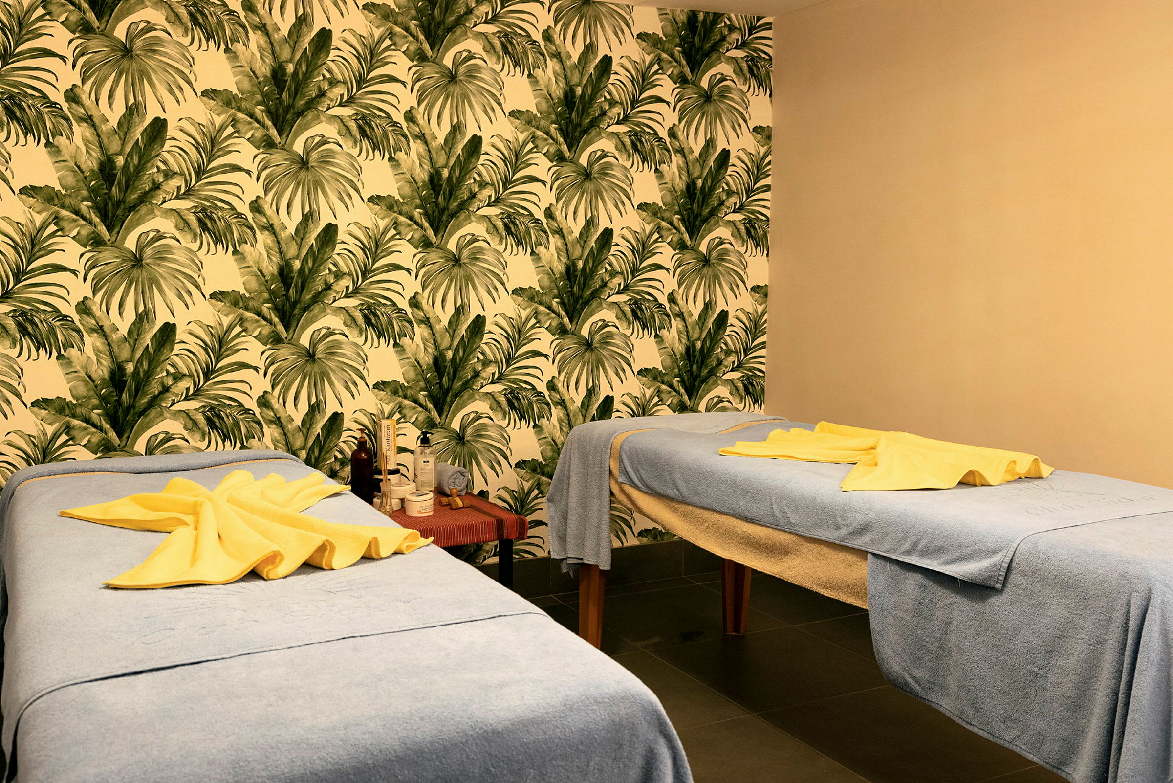 Två massagebänkar med blåa handdukar och gula handdukar ovanpå, i ett rum med tropiskt lövmotiv på väggen. (AI-genererad text)