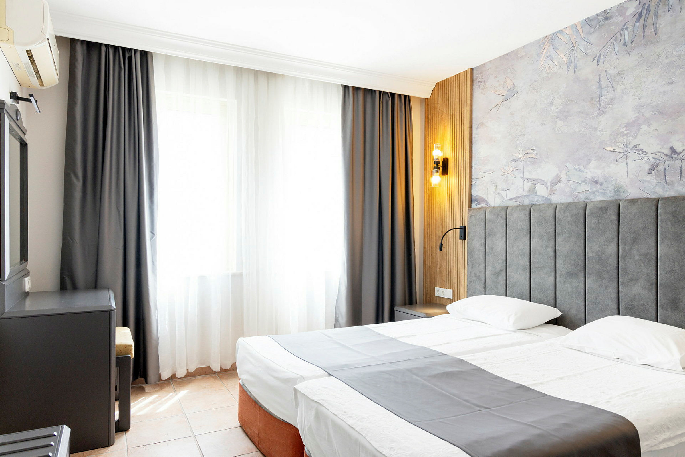 Ett hotellrum med en dubbelsäng, grått överkast, fönster med ljusa gardiner och väggpanel med dekorativt mönster. (AI-genererad text)