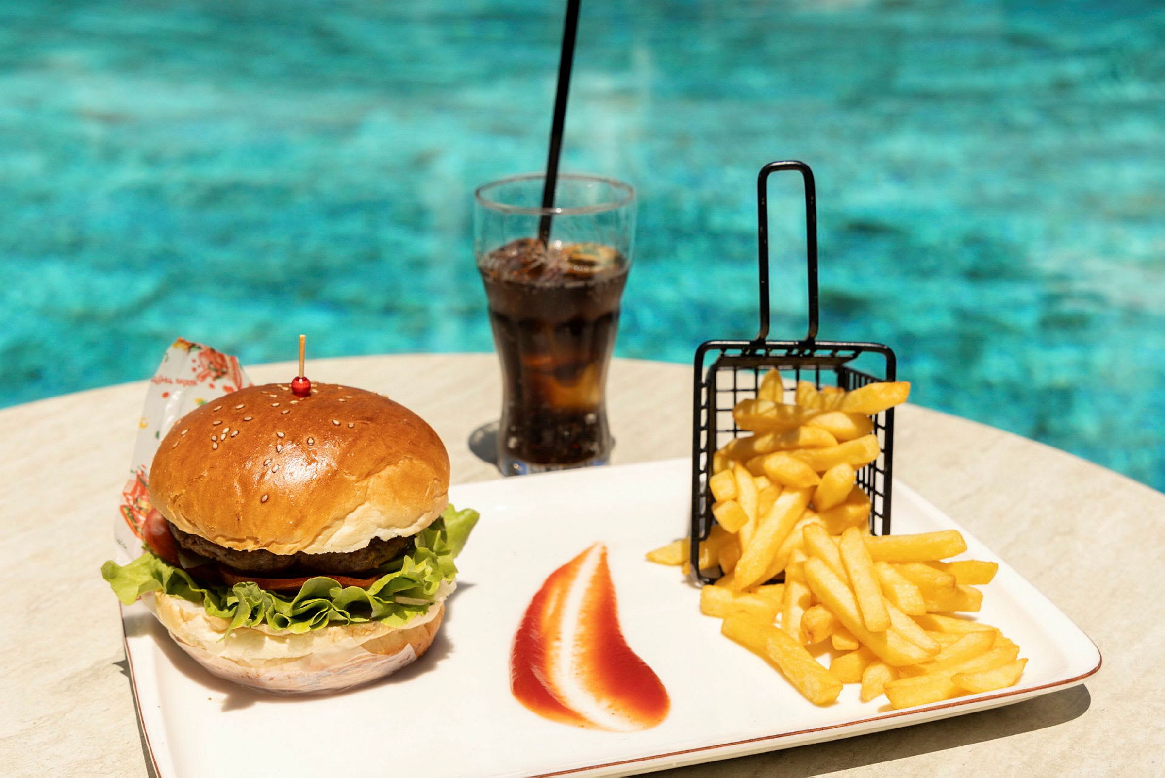 En hamburgare, pommes frites och en glas läsk på ett bord vid en pool. (AI-genererad text)