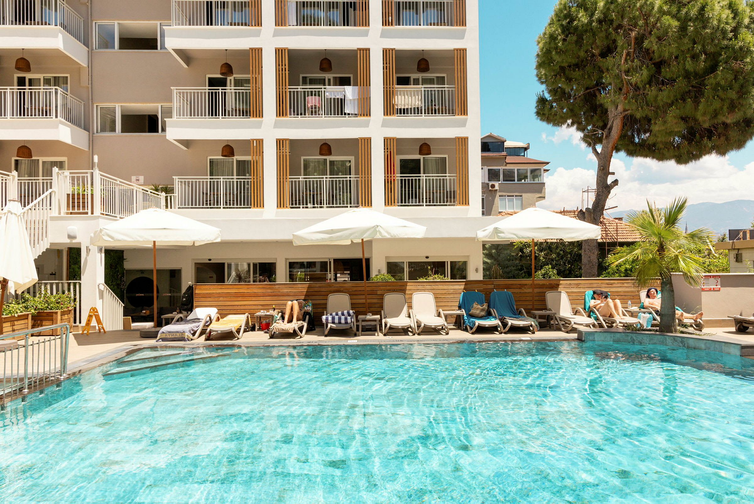 En pool med solstolar och parasoll framför en byggnad med flera våningar. (AI-genererad text)